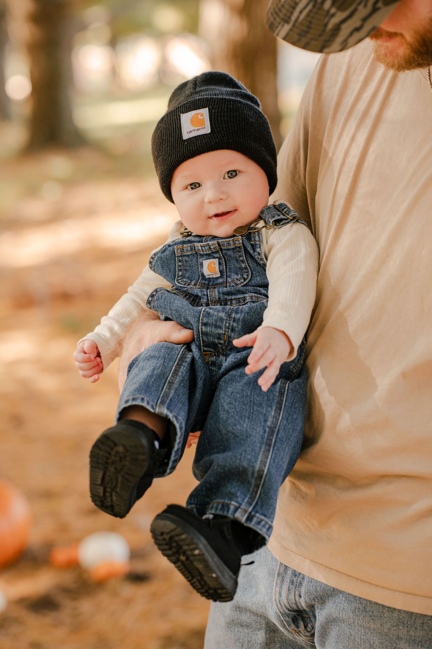 Smiling baby in a black Carhartt beanie and denim overalls held by an adult in a tan shirt outdoors near pumpkins on an autumn day.