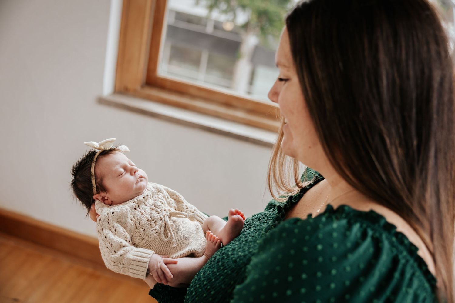 Mother in a green dress cradles her newborn baby in a cream knit outfit and bow beside a sunny window.