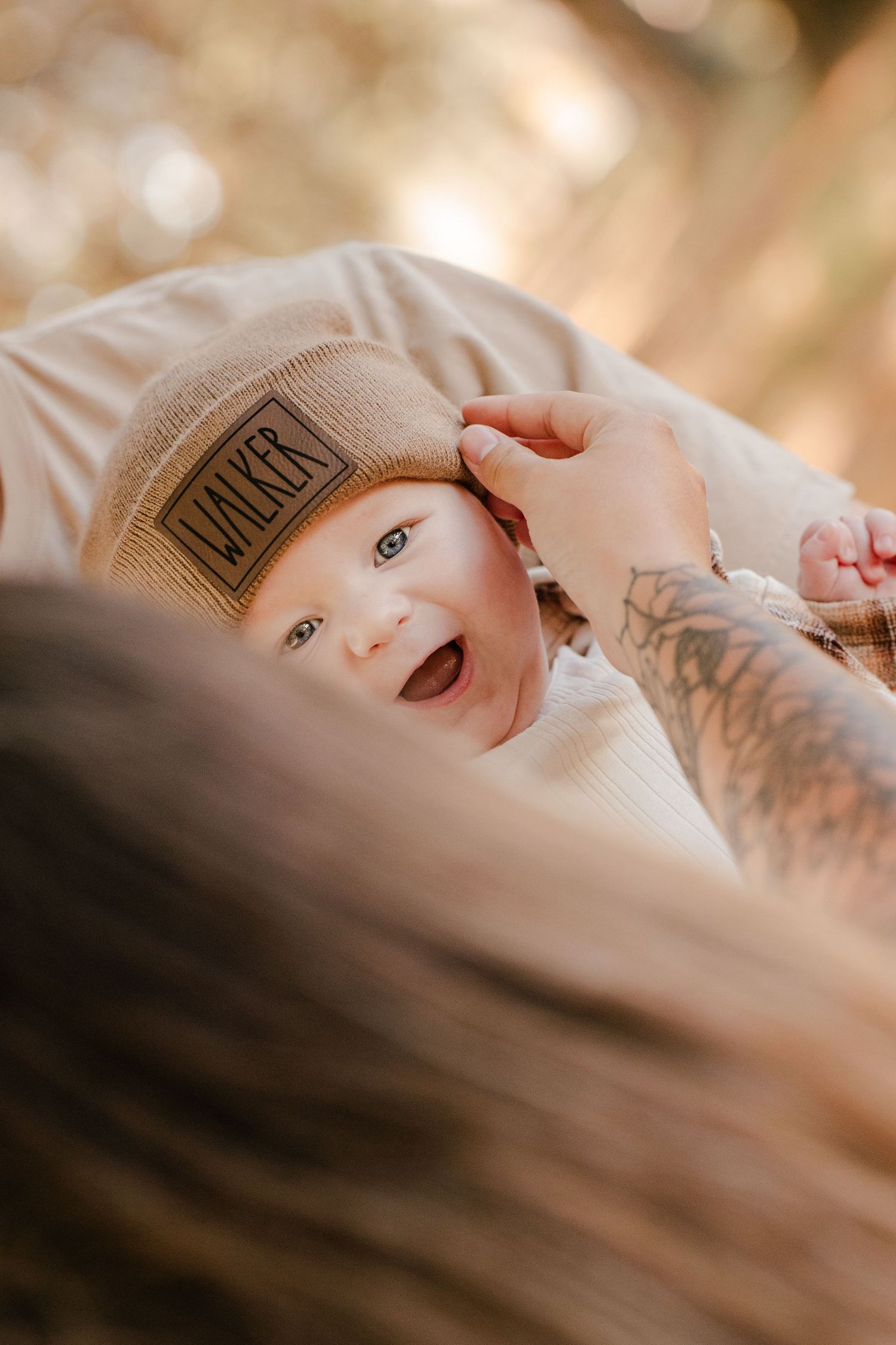 Close-up of a laughing baby in a WALKER leather-patch beanie as a tattooed parent's hand gently touches the hat.