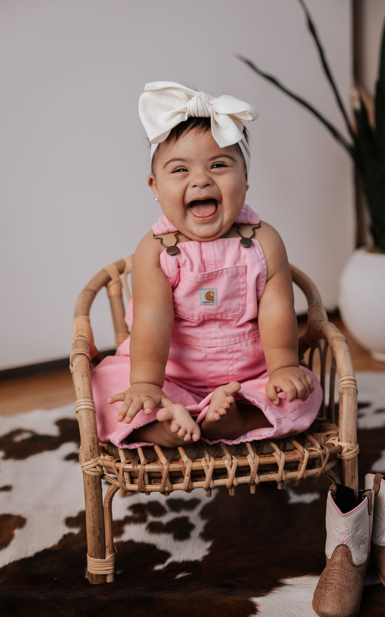 Joyful baby in pink overalls and a white bow giggles in a rattan chair, with bare feet up and cowboy boots beside her.