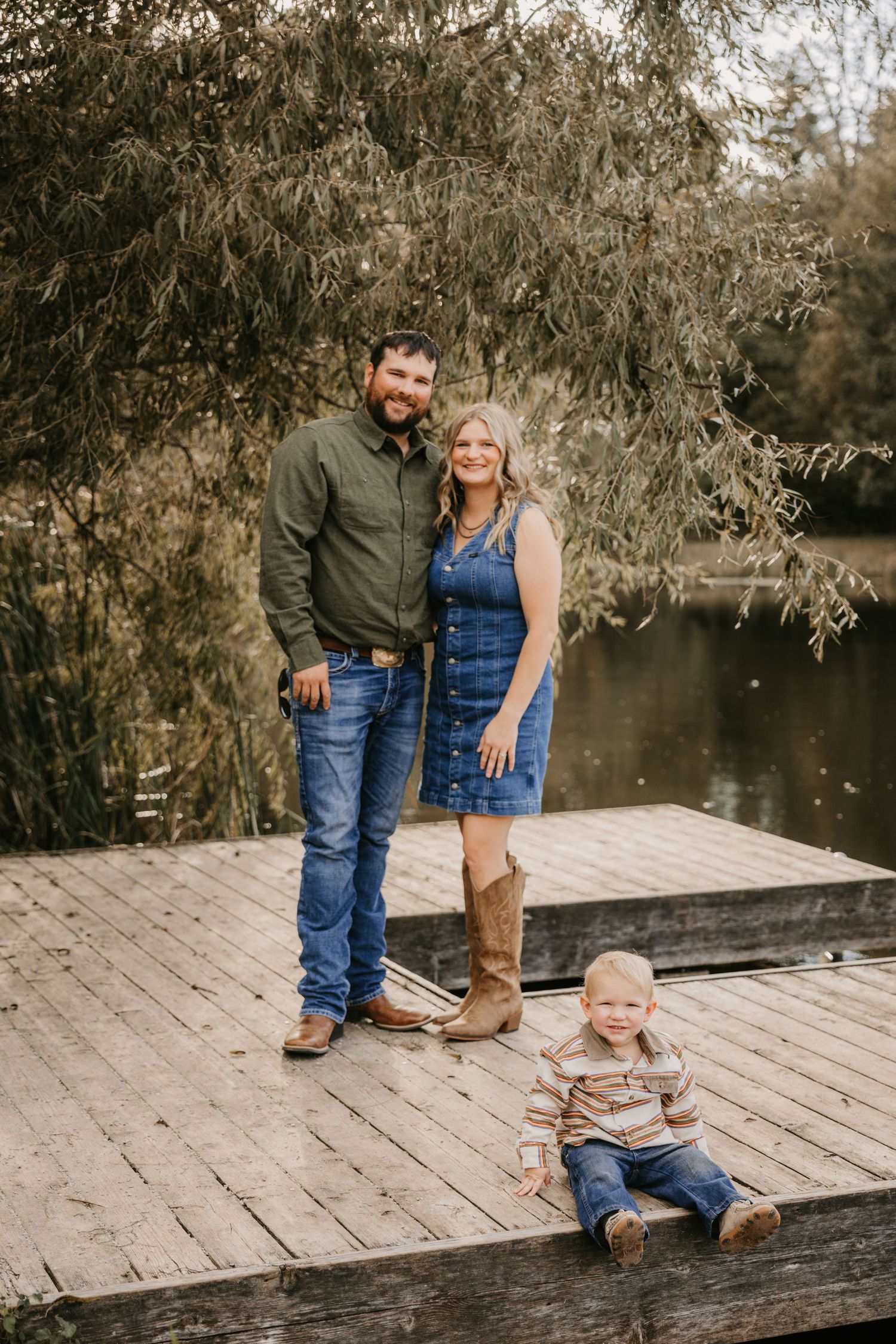 Family of three in western attire poses on a wooden lake dock beneath willow branches, with the toddler seated in front.