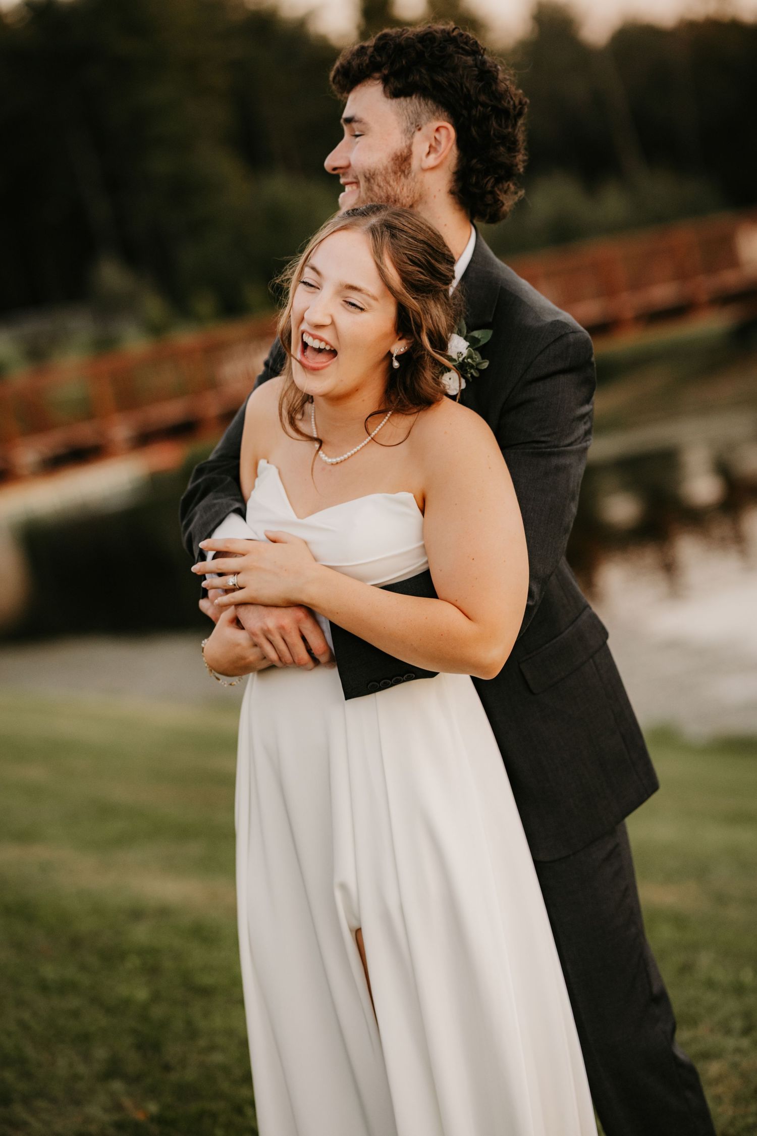 Laughing bride is hugged from behind by her groom as they celebrate on a grassy lakeside with a wooden bridge behind them.