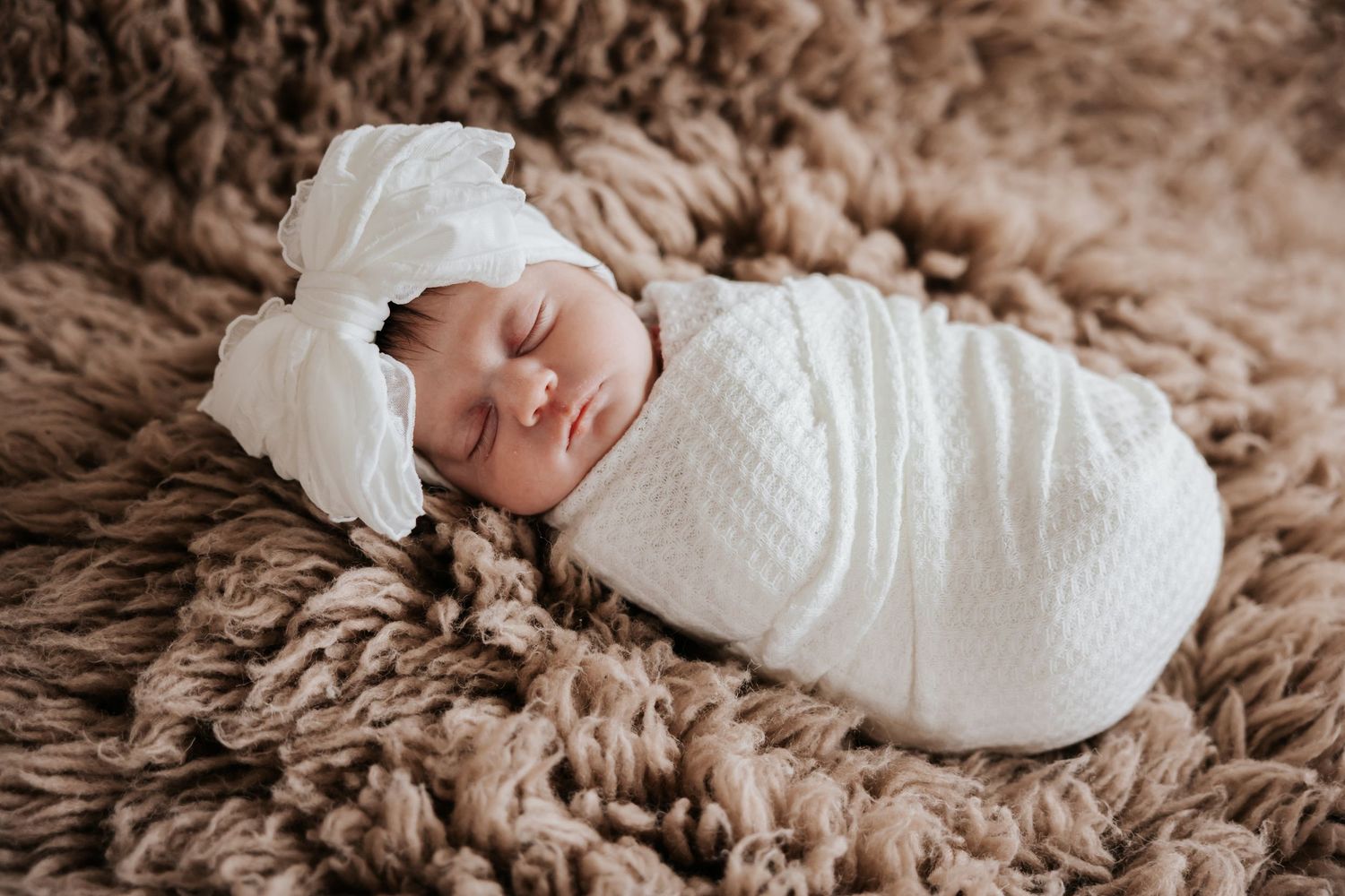 Newborn baby girl sleeps curled up in a cream knit swaddle with a soft white bow headband on a brown fluffy blanket.