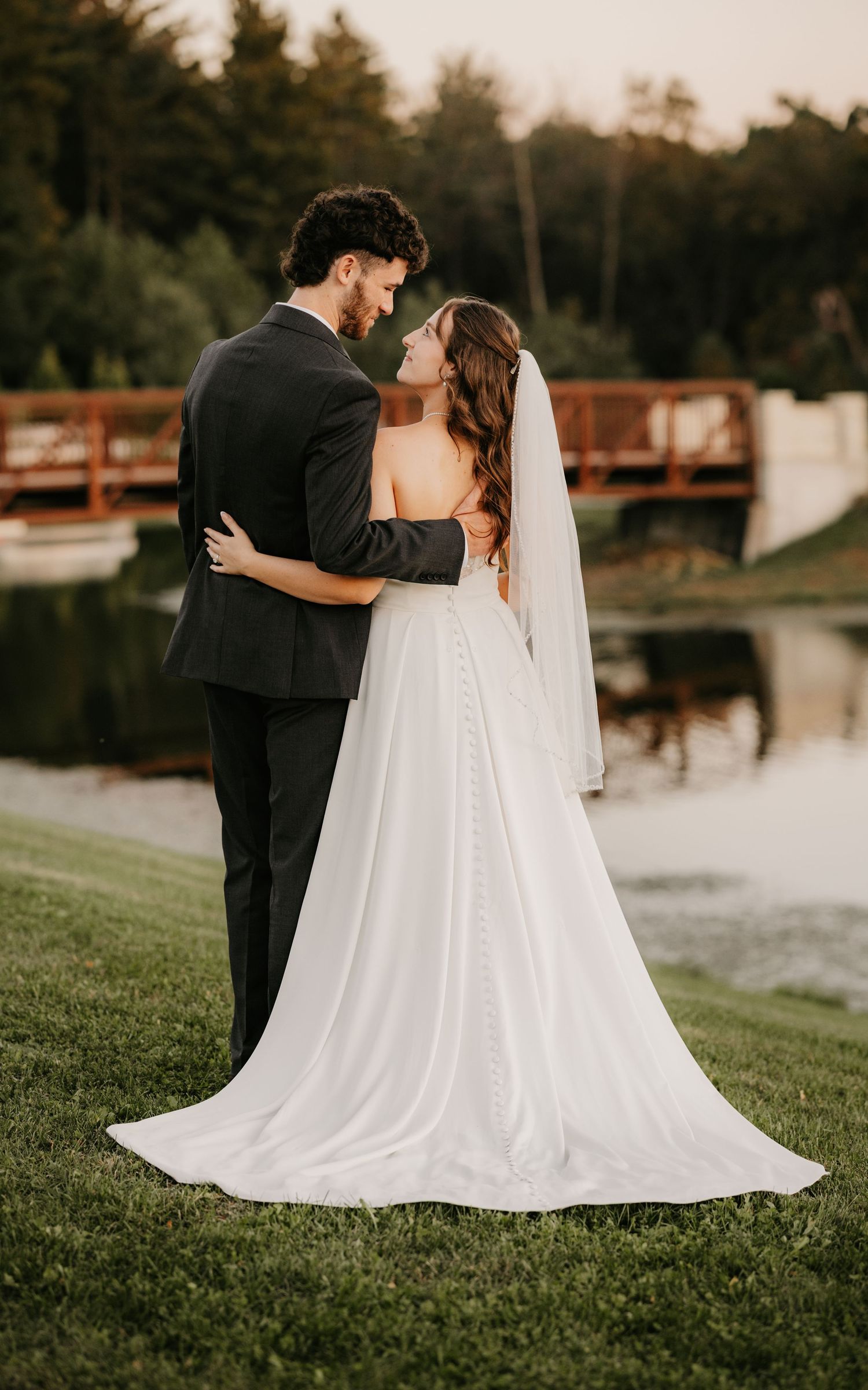 Bride in a long white gown and veil gazes back at her groom as they stand together on a lakeside lawn near a wooden bridge.