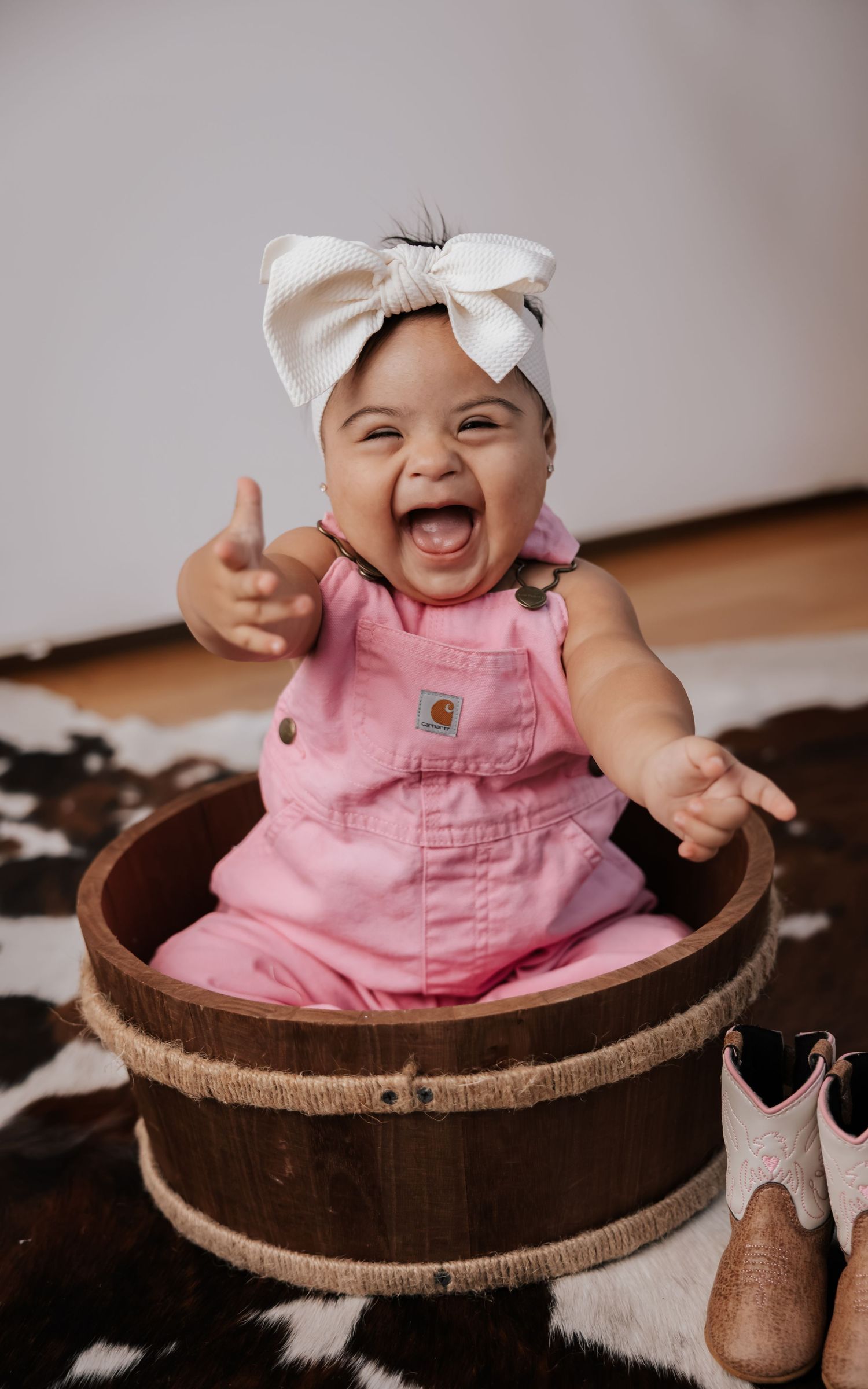 Laughing baby in pink overalls and a cream bow headband sits in a wooden barrel beside tiny cowboy boots on a cowhide rug.