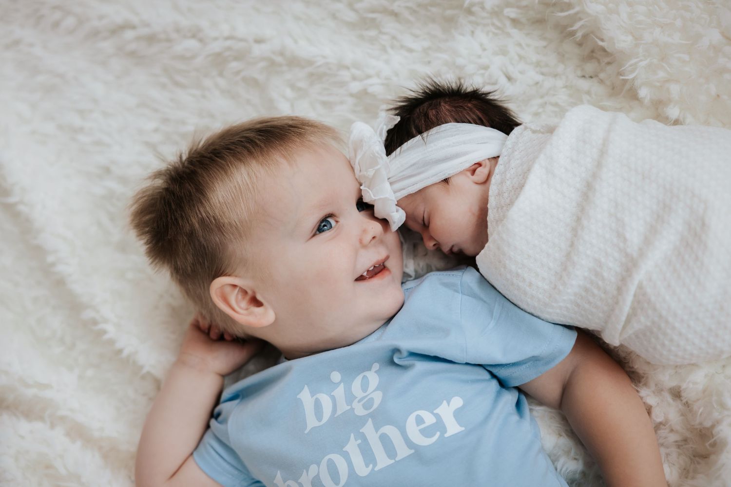 Smiling toddler in a big brother shirt lies on a fluffy white blanket beside his sleeping newborn sister in a white bow headband.