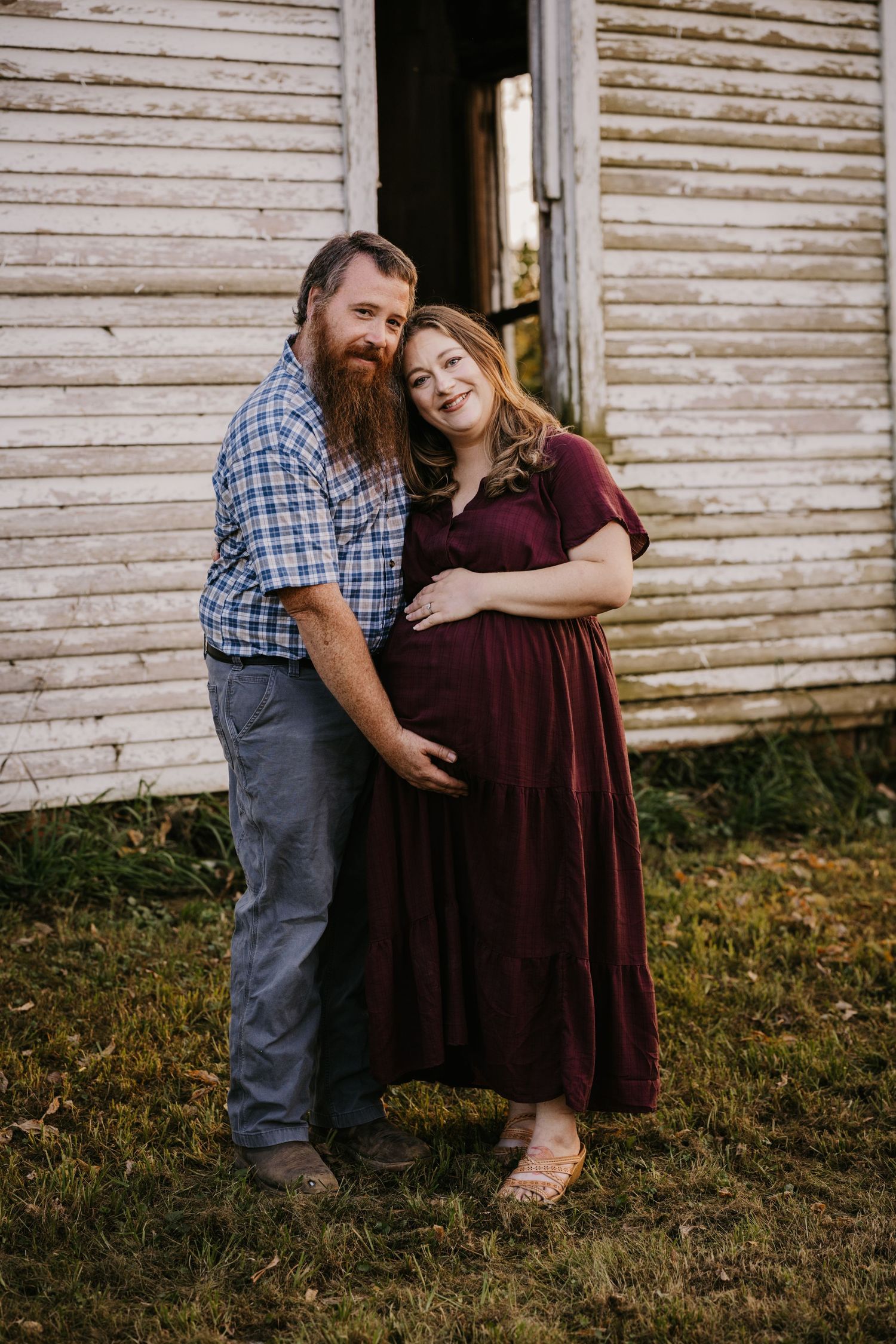 Happy couple poses for a maternity portrait, the husband's hand on his wife's baby bump, in front of a rustic white wooden barn.