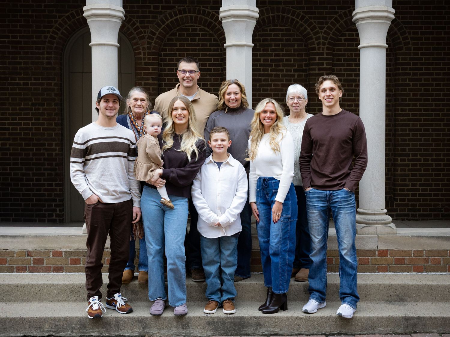 Smiling extended family of all ages standing together on brick steps in front of white columns and a dark brick wall.