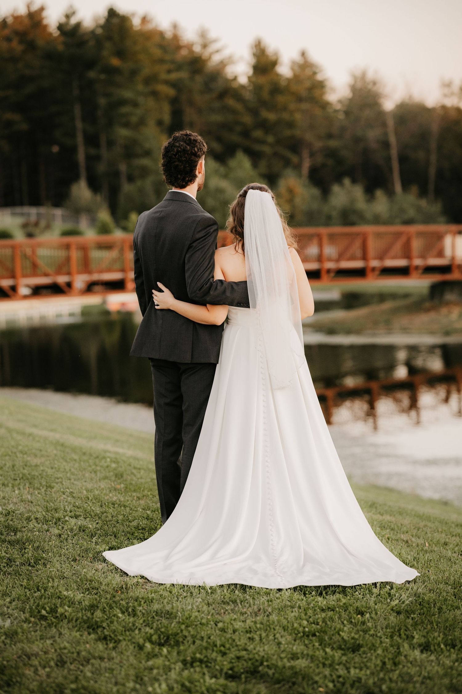 Newlyweds embrace and look out over a peaceful pond toward a wooden bridge, the bride's long veil trailing on the grass.