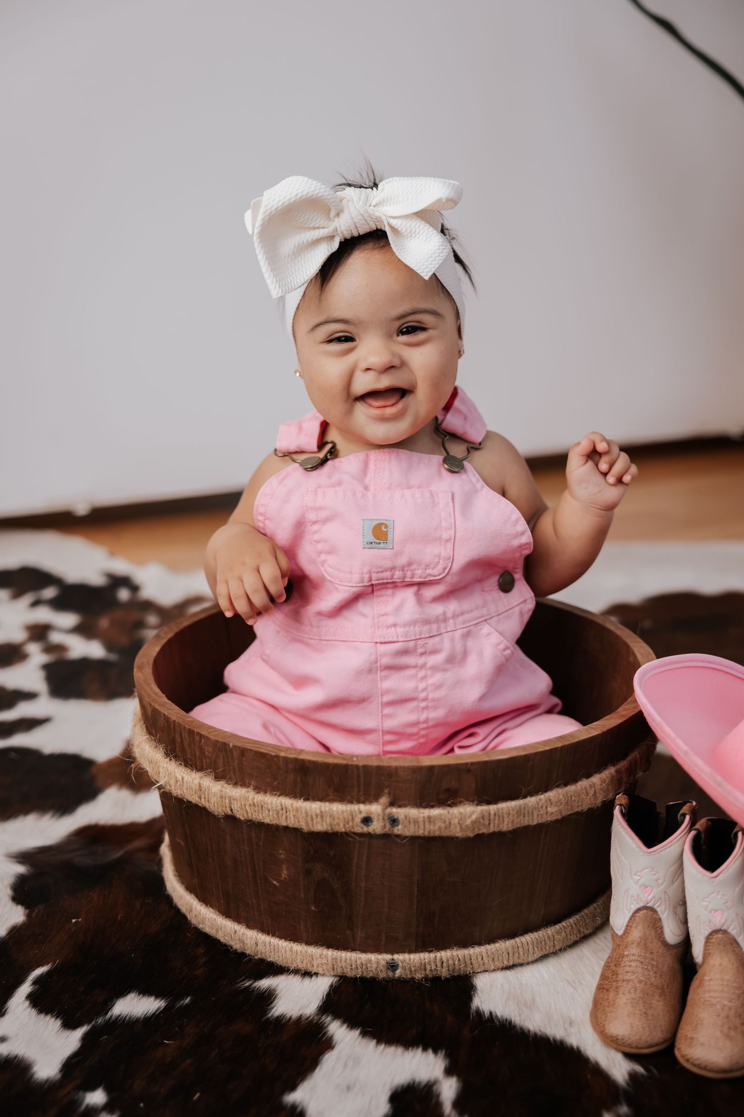 Laughing baby girl with a white bow headband and pink overalls sits in a rustic wooden barrel beside tiny cowgirl boots.