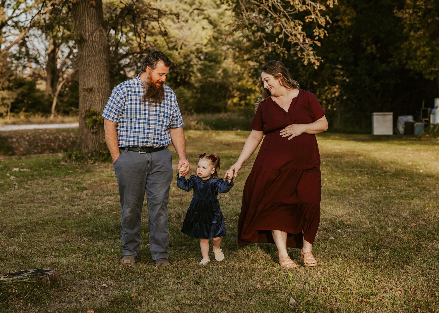 Bearded father, pregnant mother and toddler girl hold hands and walk across a grassy field during a sunny family photo session.