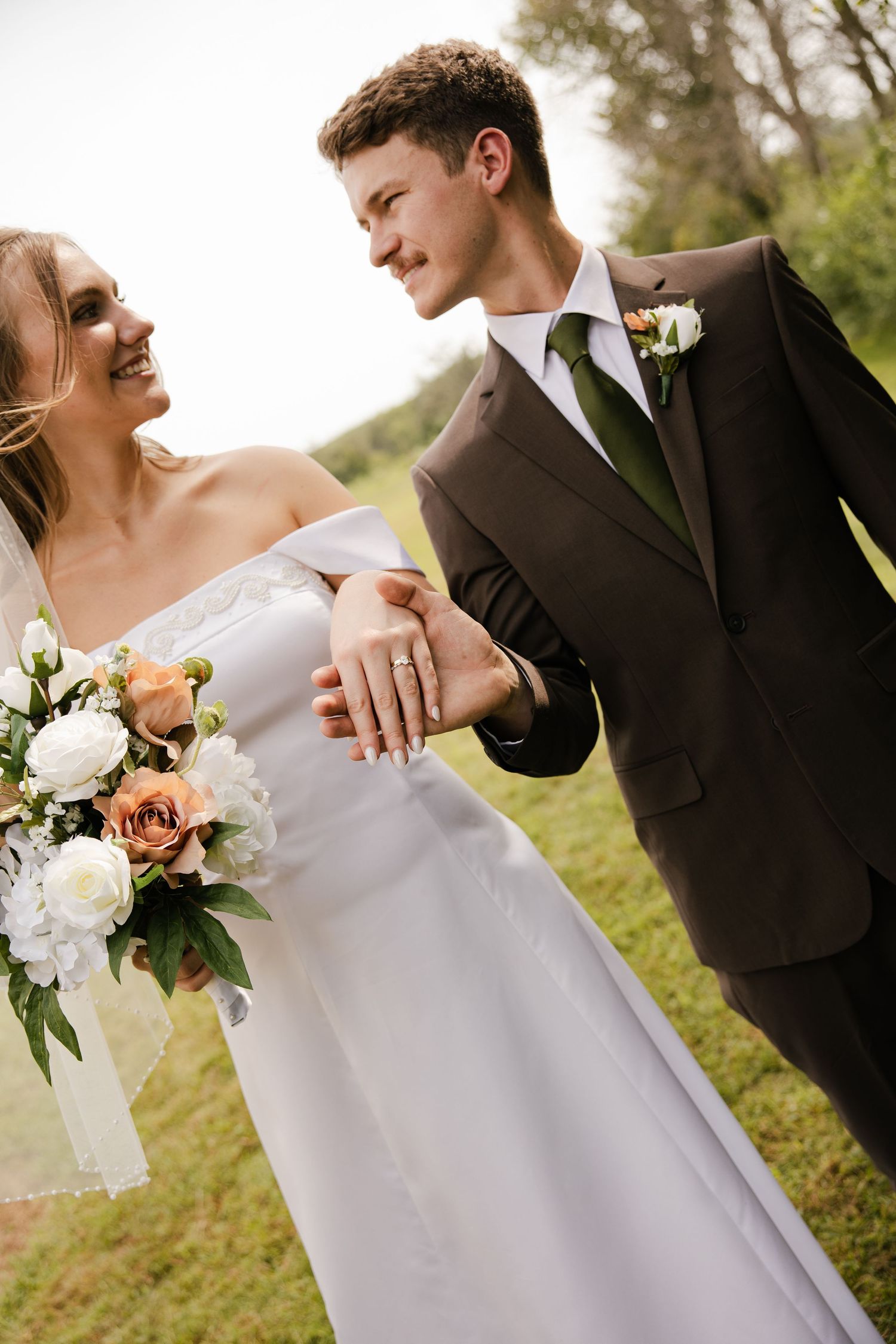 A groom in a brown suit and green tie smiles at his laughing bride as they walk hand in hand, her bouquet of white and peach roses nearby.