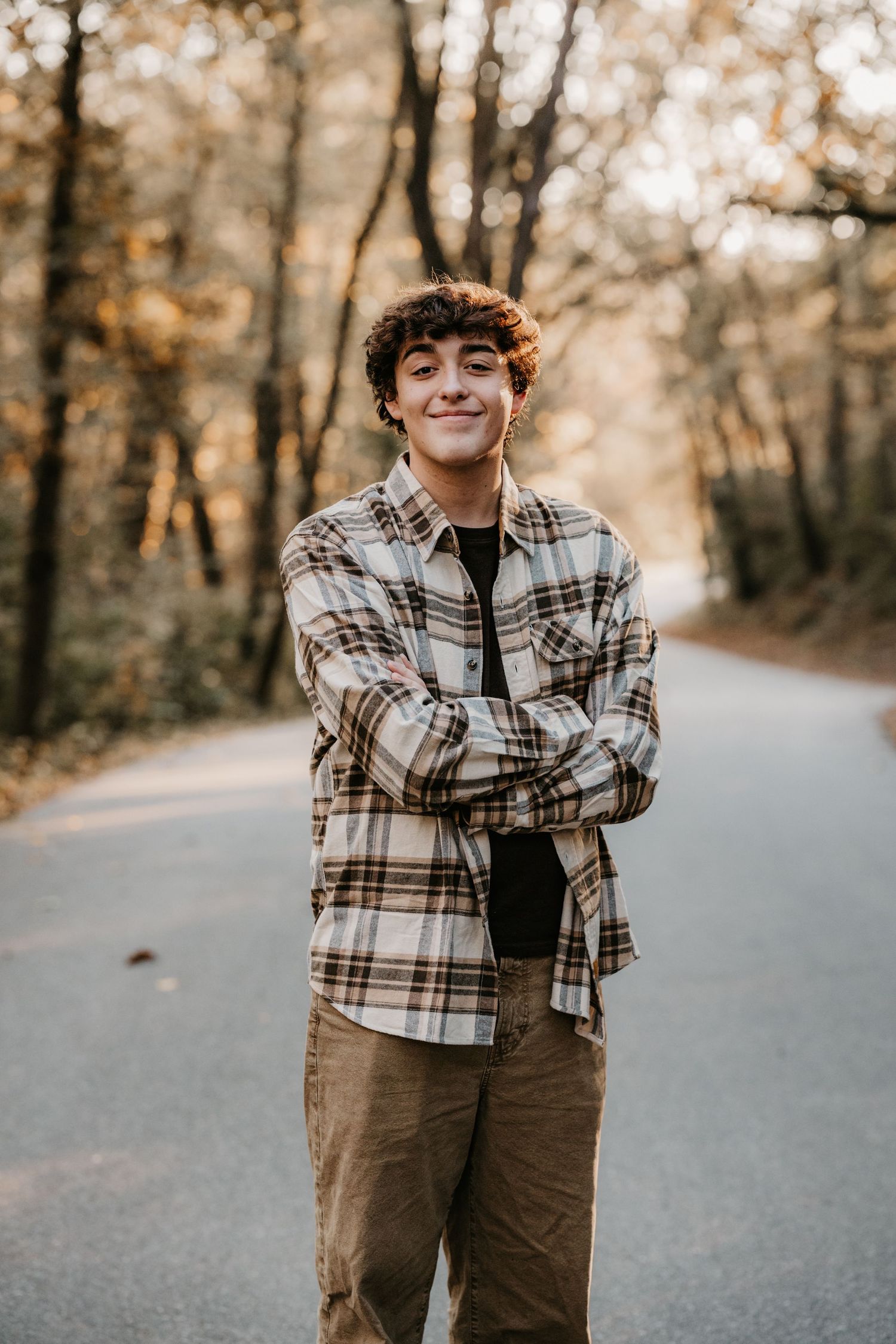 Smiling young man in a plaid flannel shirt and khaki pants poses with crossed arms on a quiet road surrounded by fall trees.