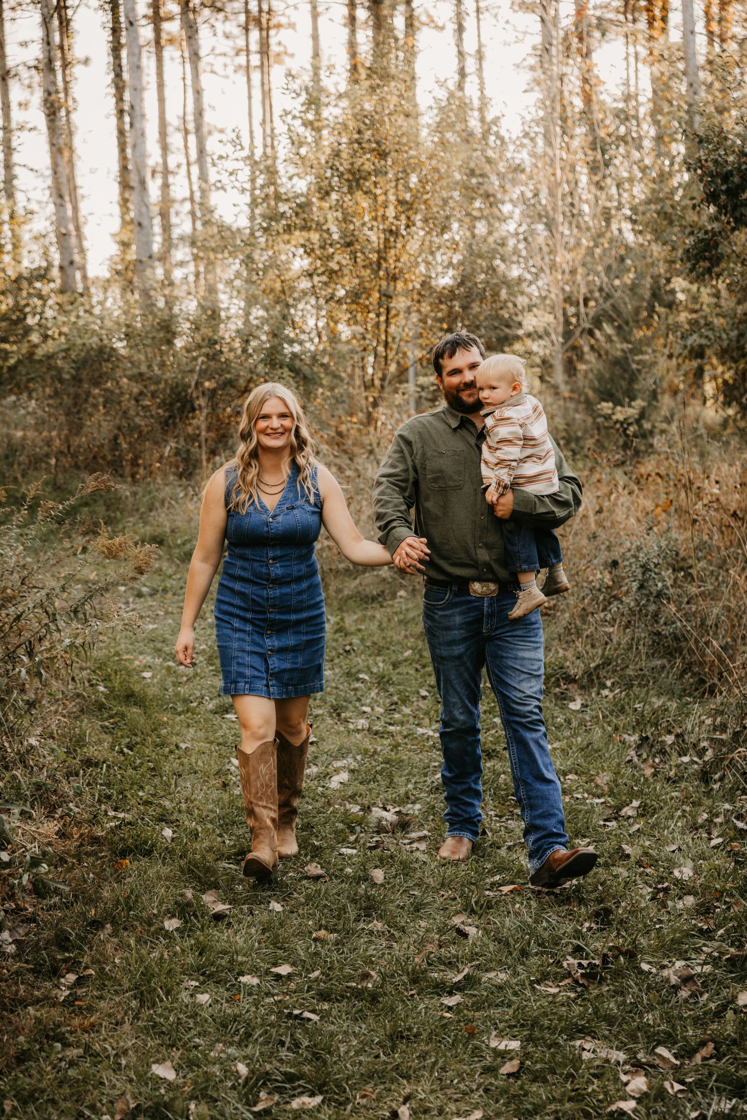 A smiling couple walks hand in hand through an autumn forest while the man carries a toddler in a striped sweater.