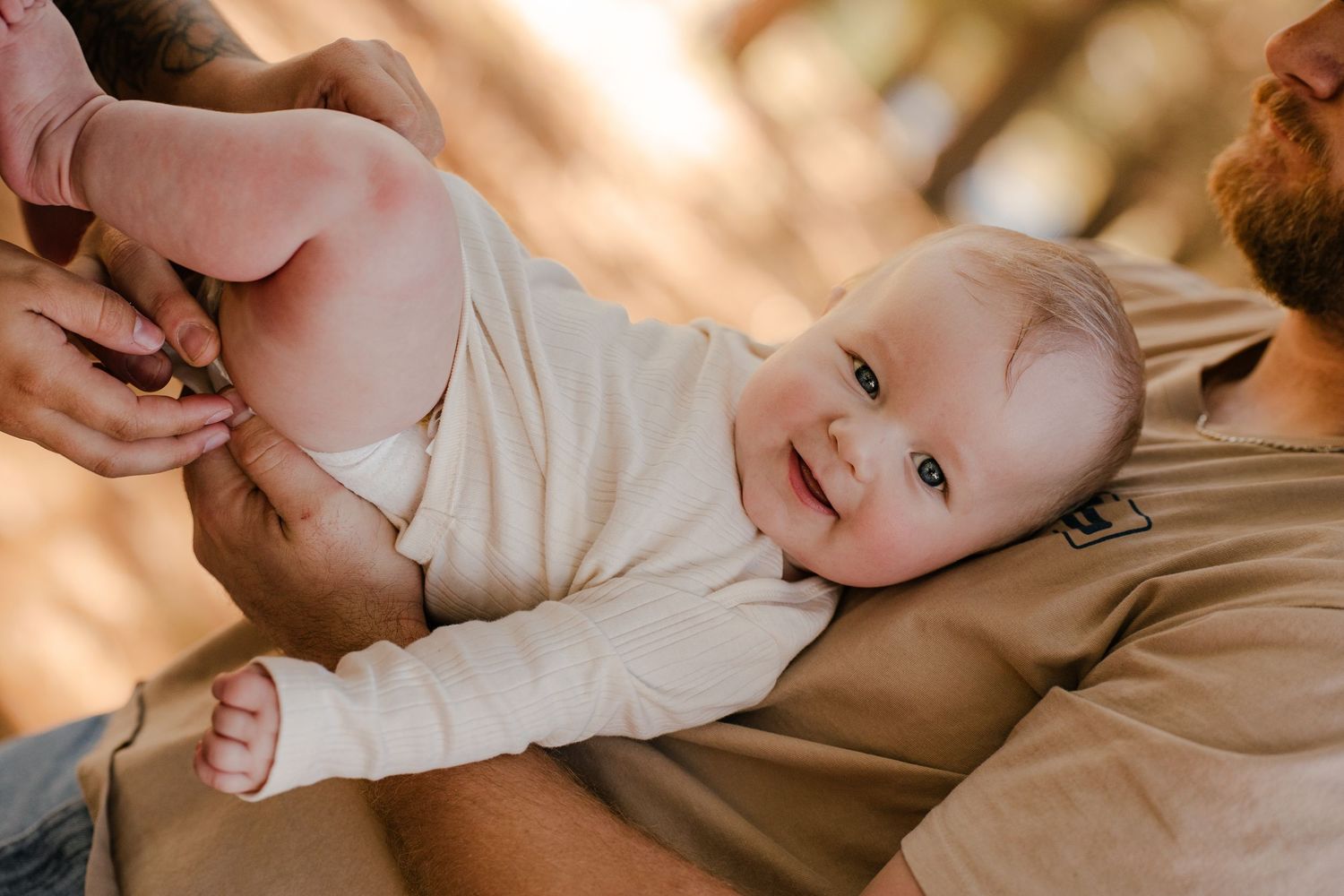 Smiling baby in a cream bodysuit lies on a bearded dad's chest while a parent gently holds the baby's leg in warm outdoor light.