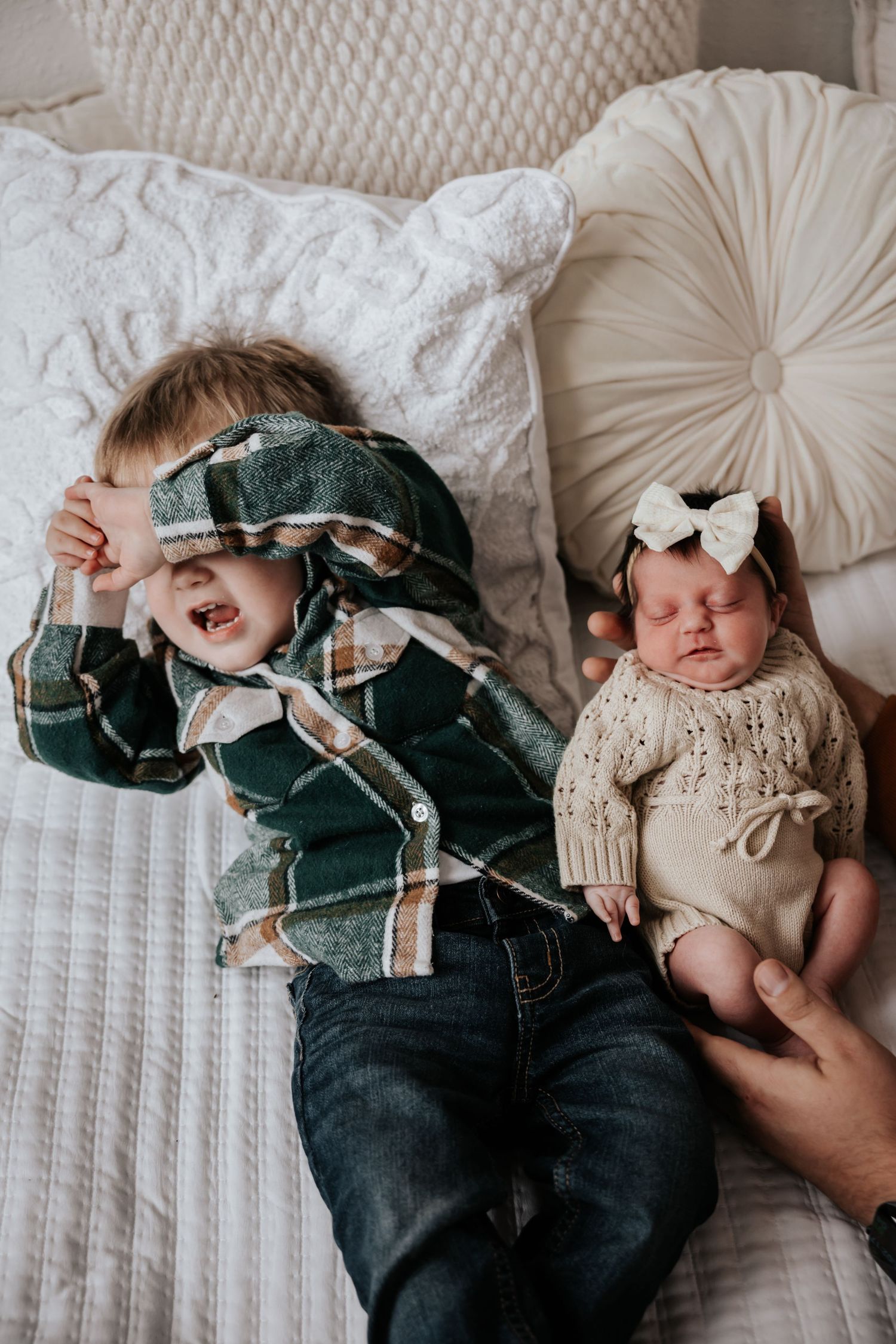 Toddler boy in a green plaid shirt covers his eyes on a bed beside his sleeping newborn sister wearing a white bow.