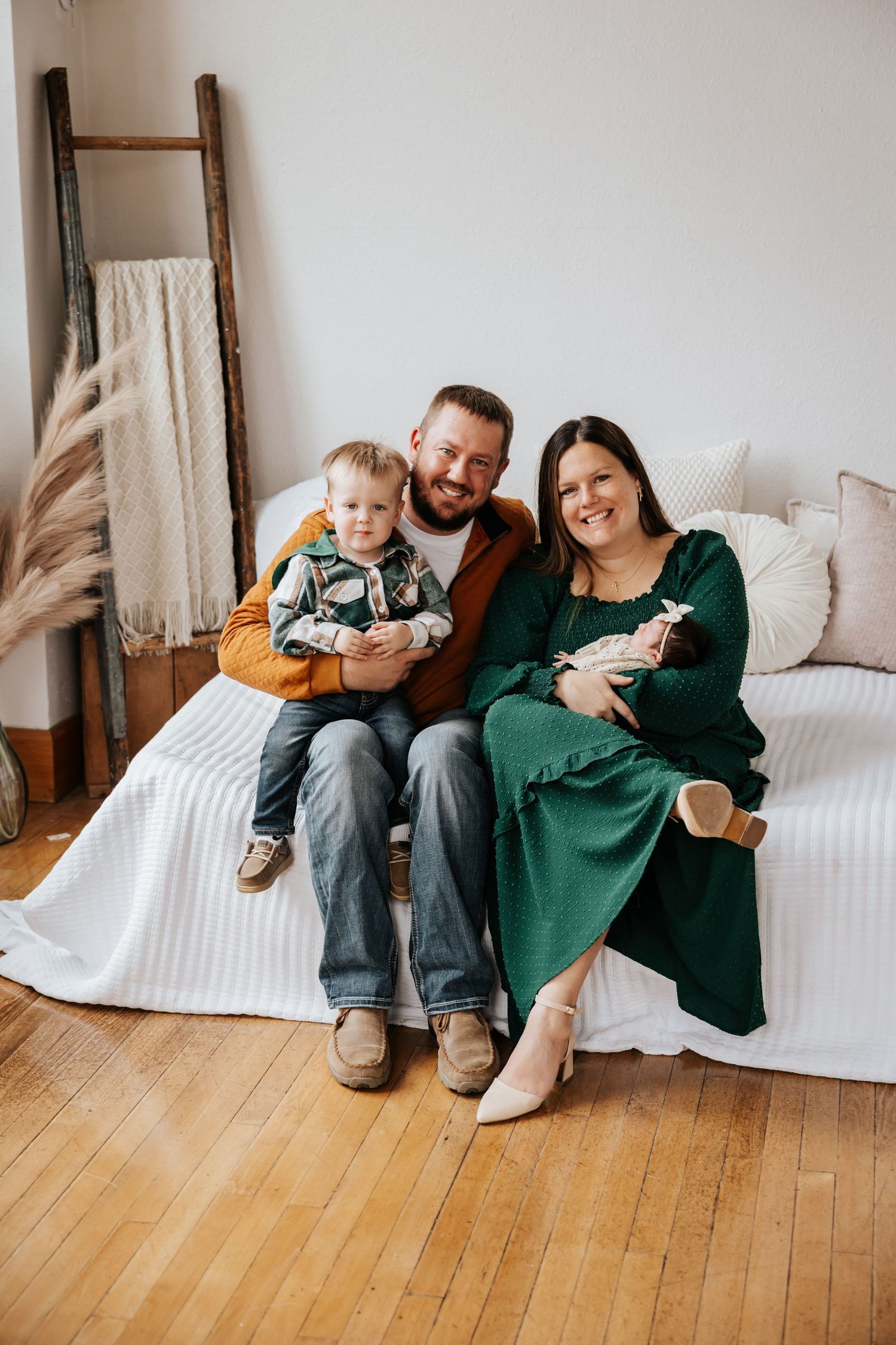 A family of four sits together on a white daybed, with a father holding a toddler and a mother cradling a sleeping newborn.