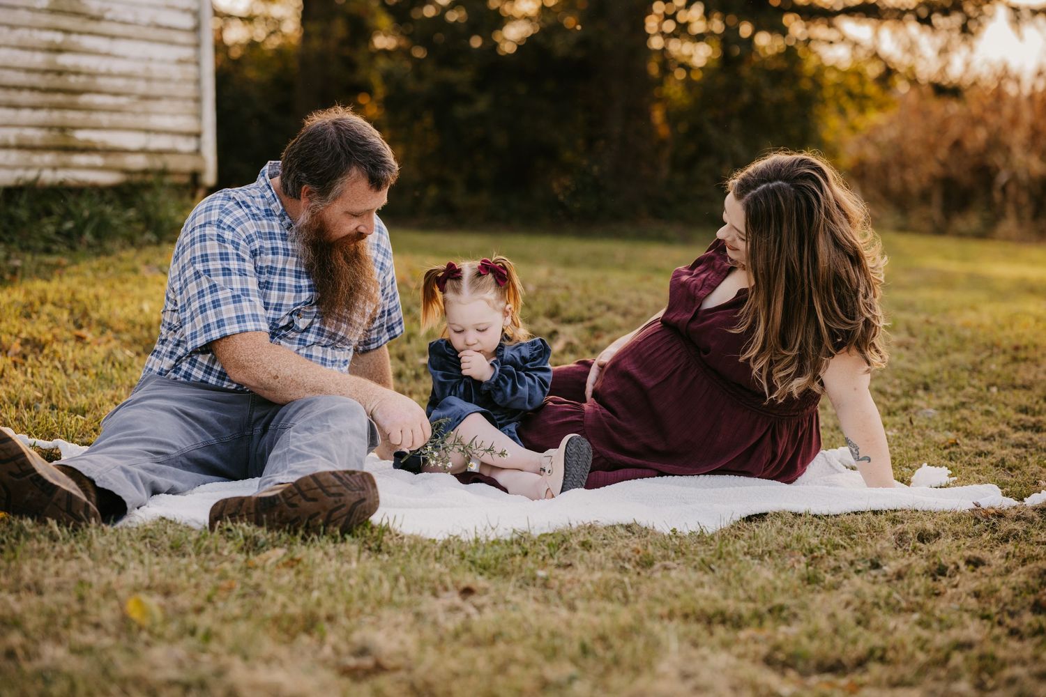 Family of three, with an expecting mother, sits on a blanket in golden-hour light as the toddler plays between her parents.