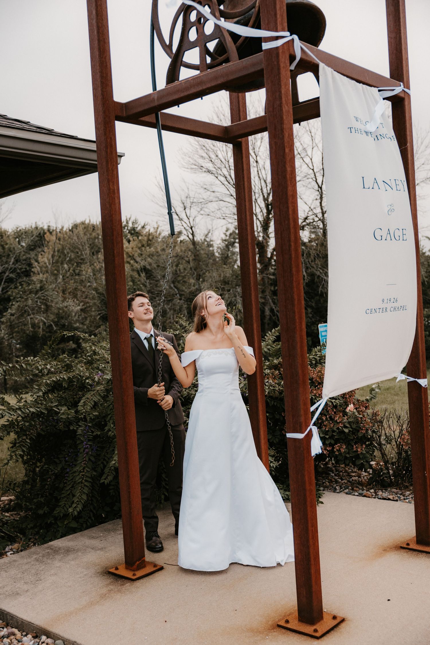 A bride in a white gown looks up at a rust-colored bell tower as a groom stands beside her, with a wedding banner for Laney and Gage.