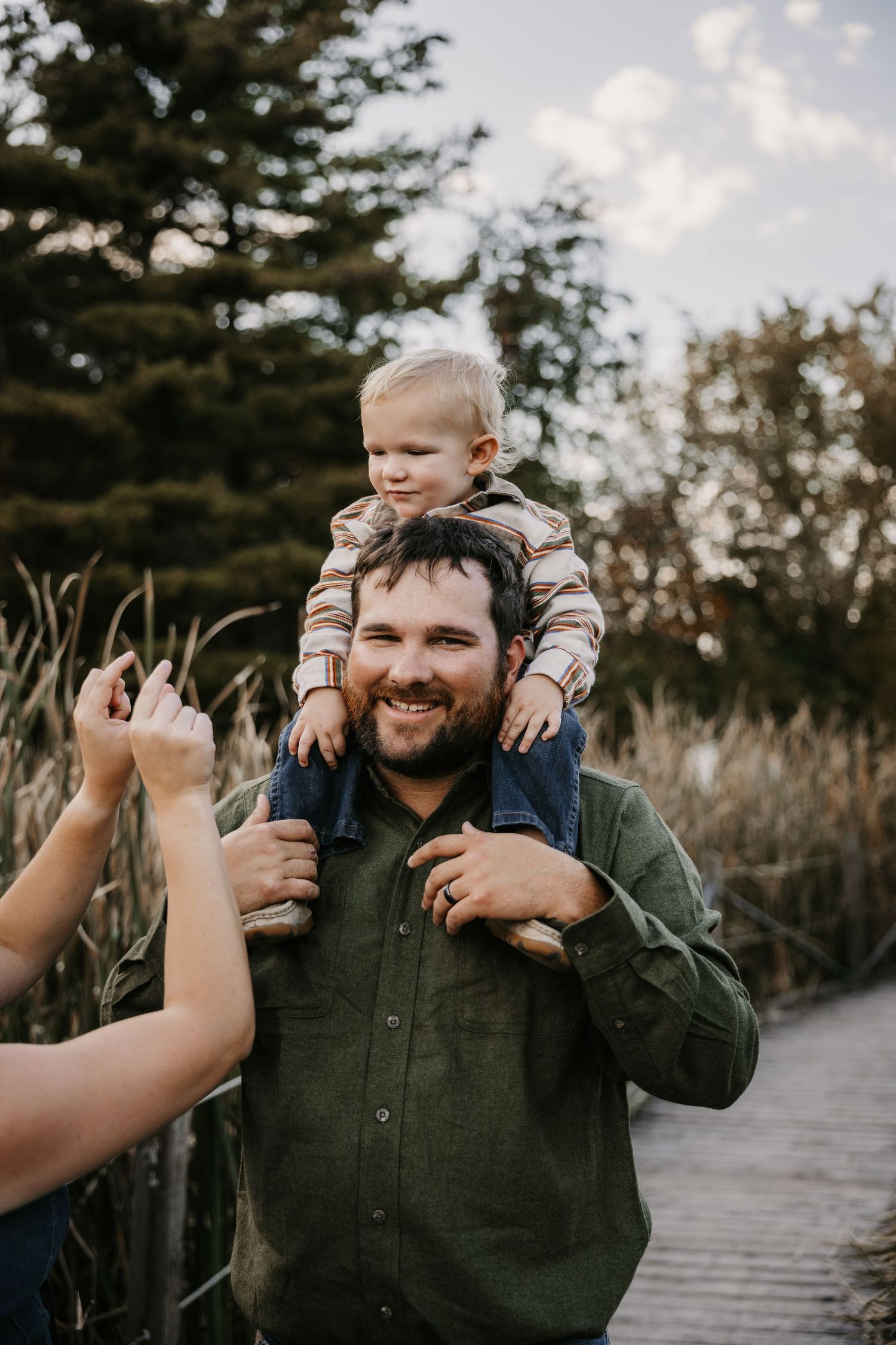 A smiling bearded man in a green shirt carries a toddler on his shoulders near tall grass and evergreen trees.