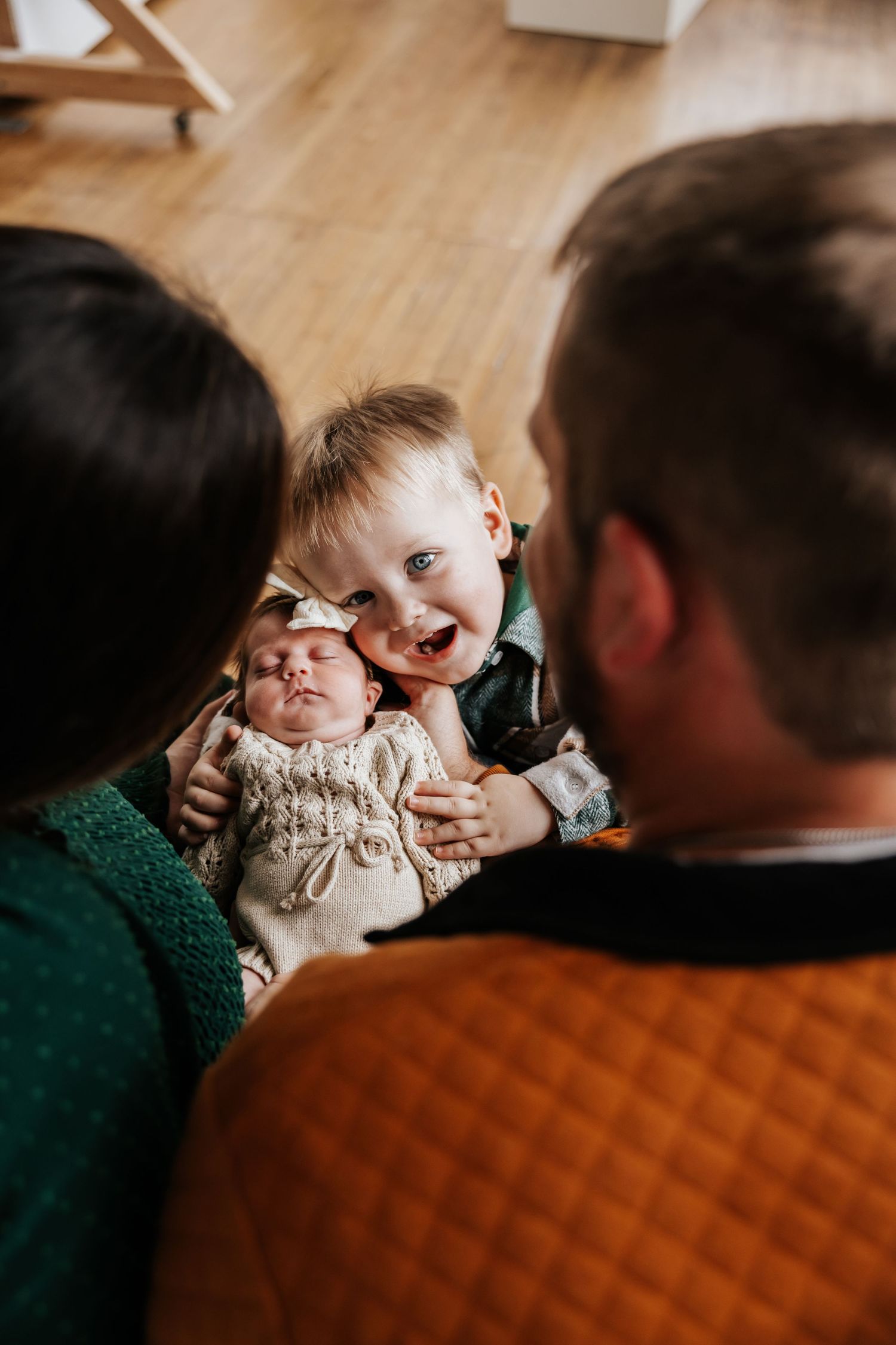A curious toddler leans in beside a sleeping newborn sibling as their parents look on in a sweet family photograph.