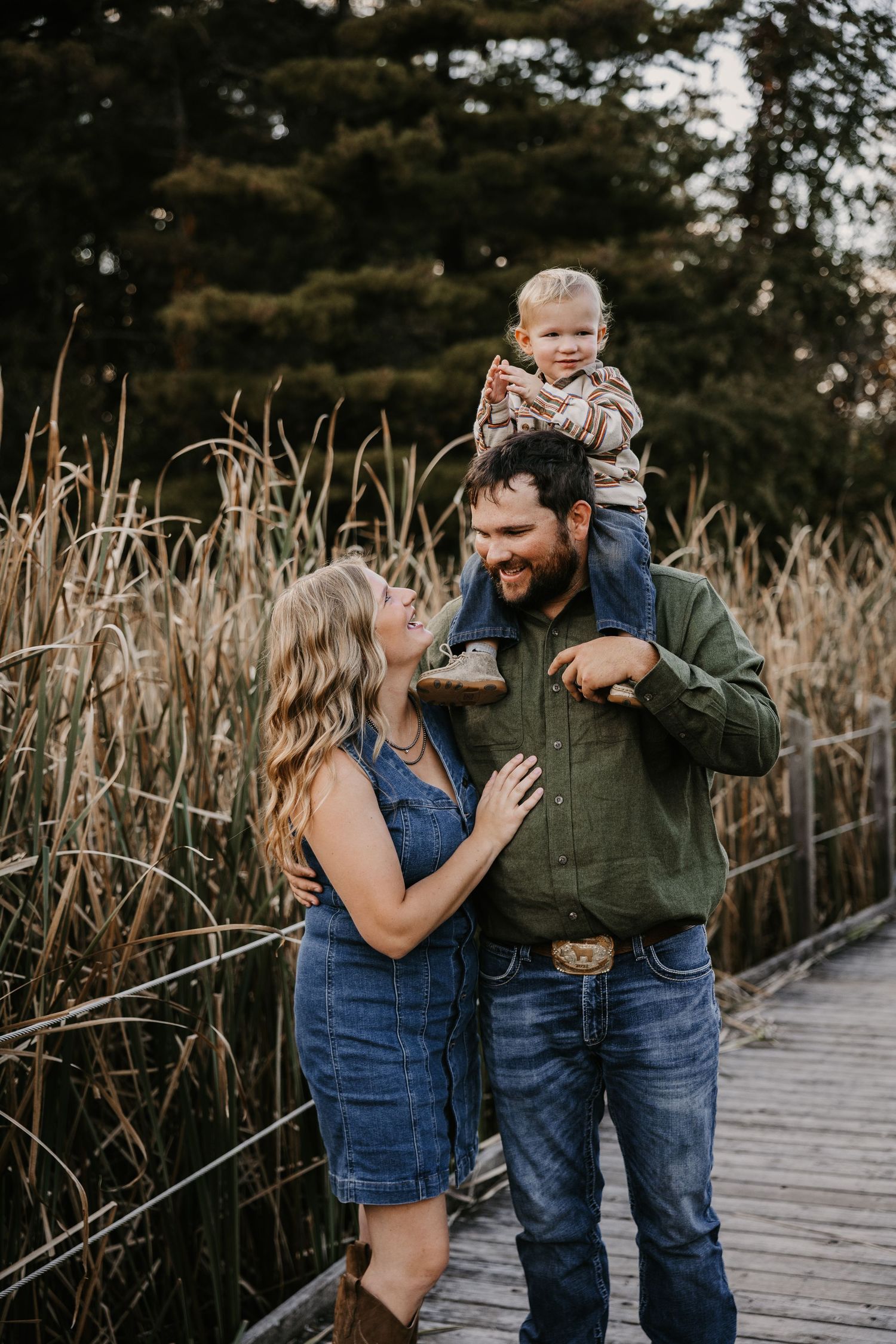 A woman gazes lovingly at her bearded husband as he carries their toddler on his shoulders on a boardwalk by tall reeds.