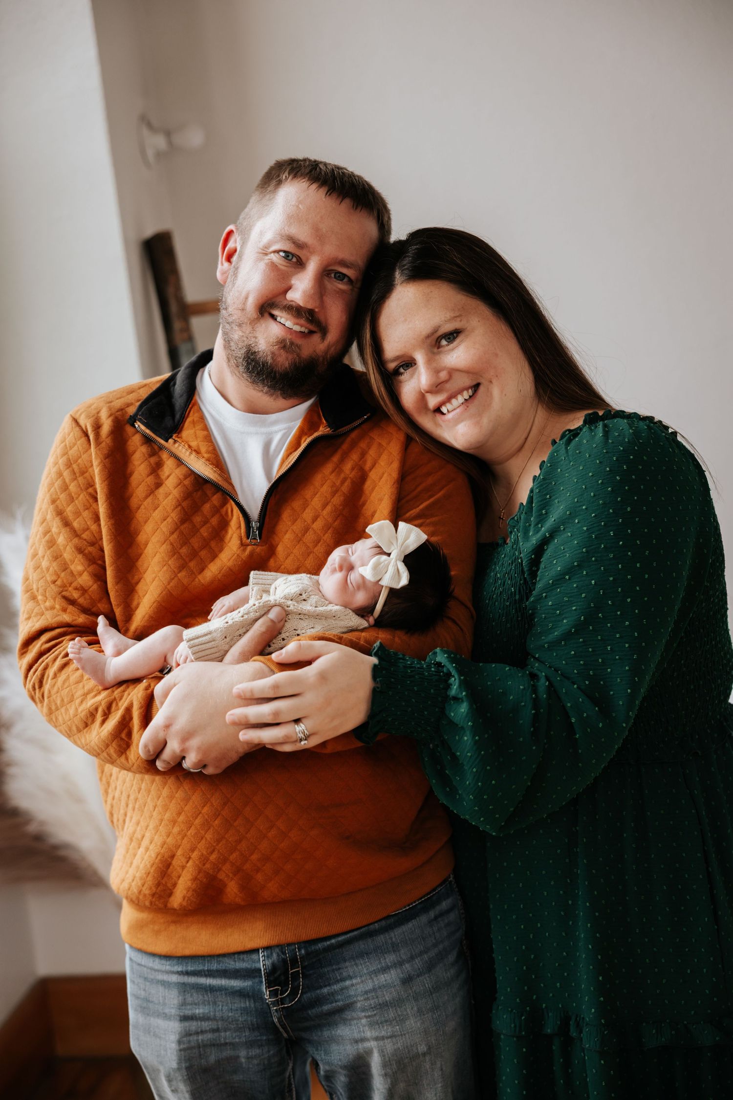 Smiling couple in an orange pullover and green dress cuddle a sleeping newborn baby in a cream outfit and bow headband.