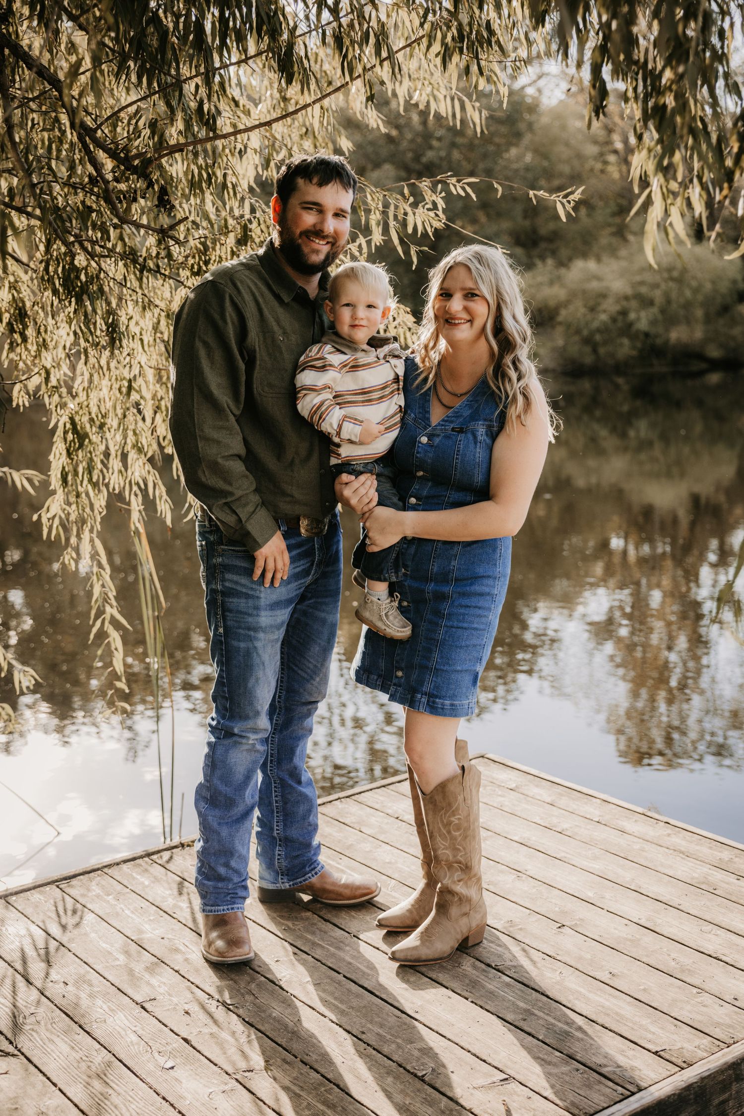 Smiling father and mother in denim and boots hold their toddler on a lakeside wooden dock under hanging willow branches.