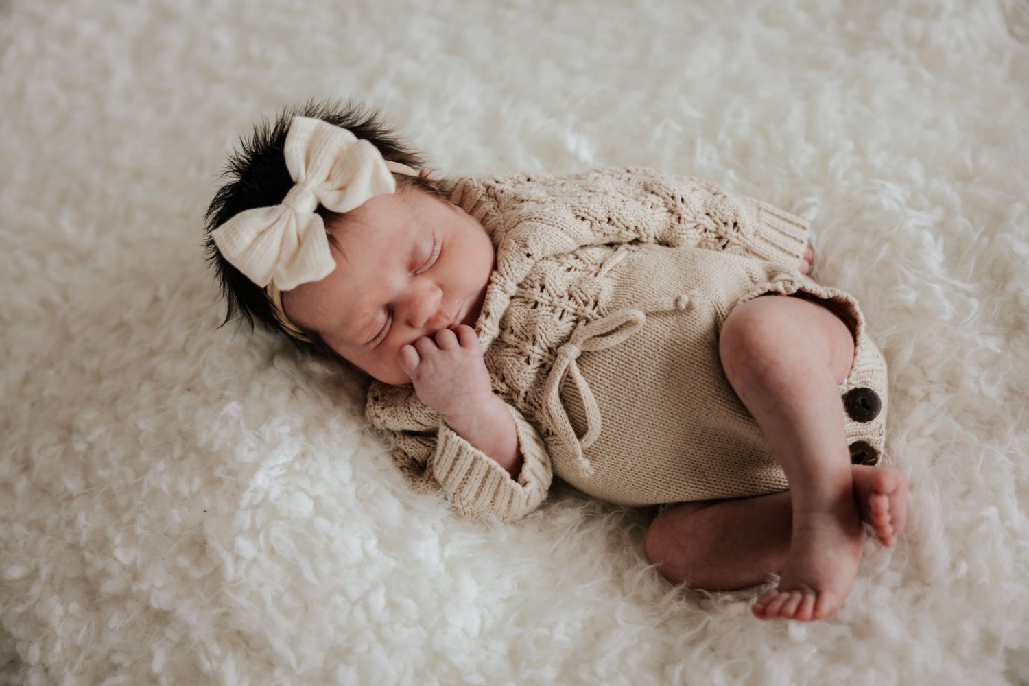 Newborn baby girl with dark hair sleeps on a white fluffy blanket wearing a cream bow headband and a knit romper with buttons.