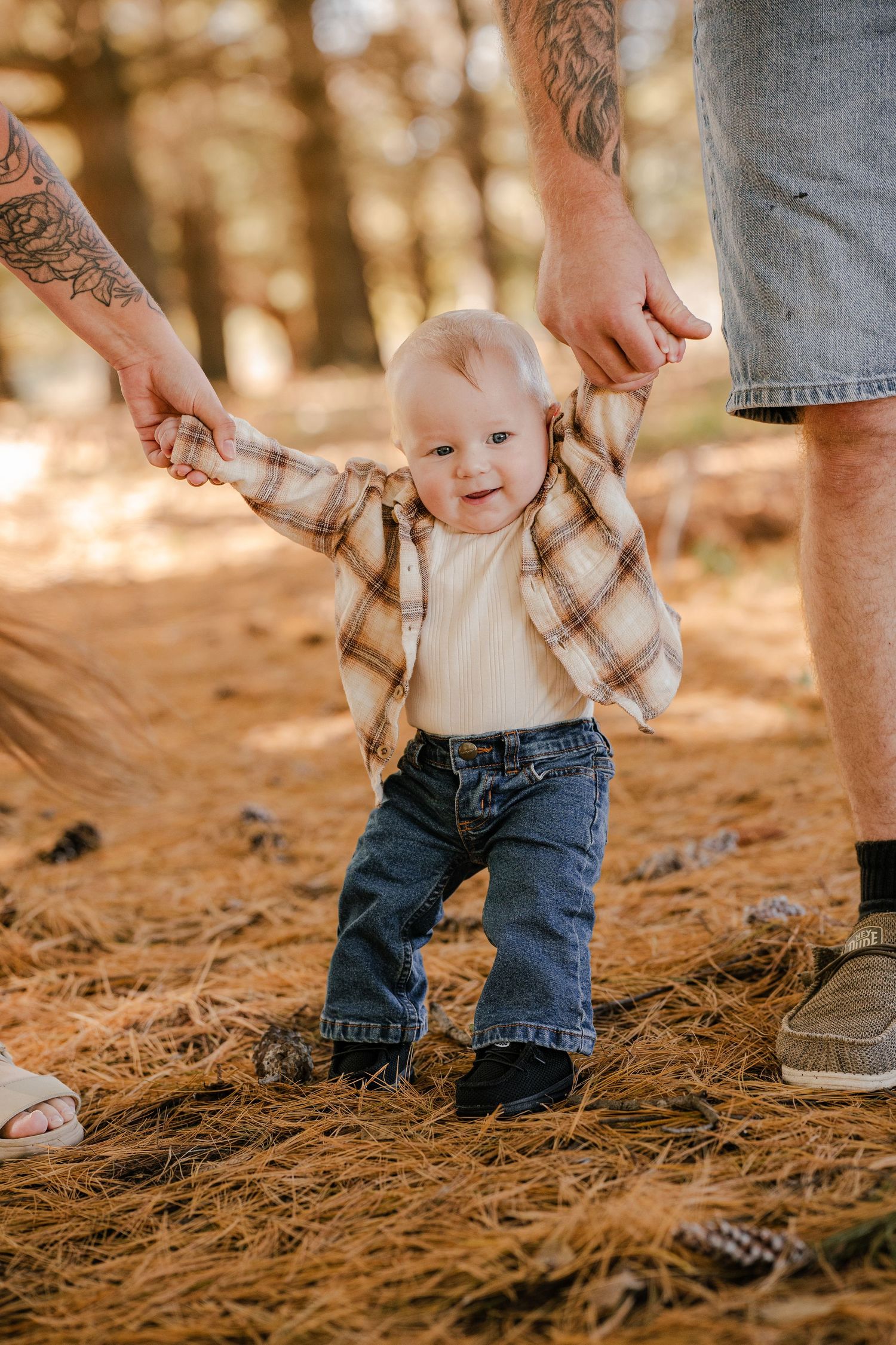 Baby in plaid flannel and jeans takes wobbly first steps on pine needles while a tattooed parent holds both hands.