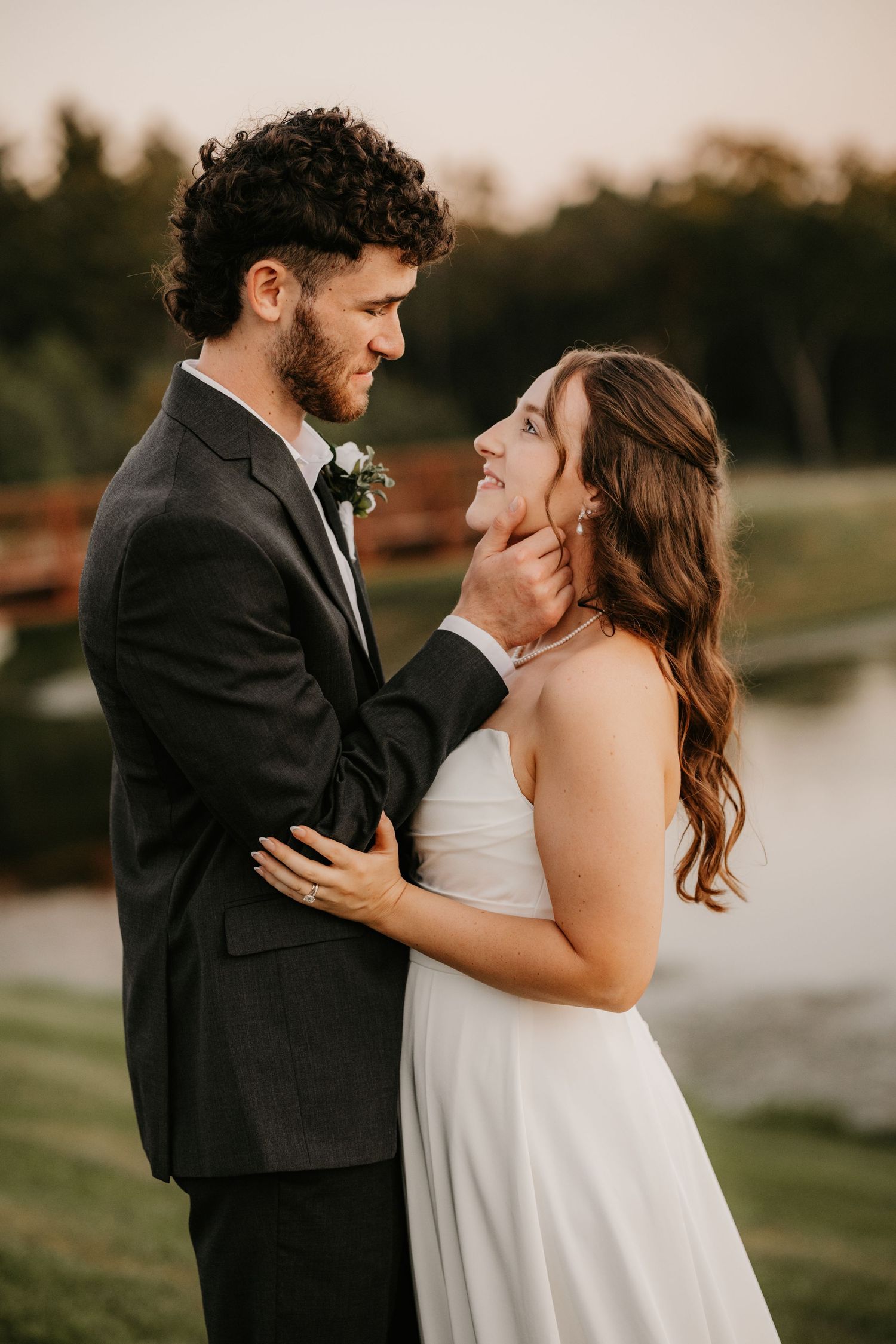 Groom with curly hair cradles his bride's chin as she smiles up at him in a white strapless gown beside a calm pond.