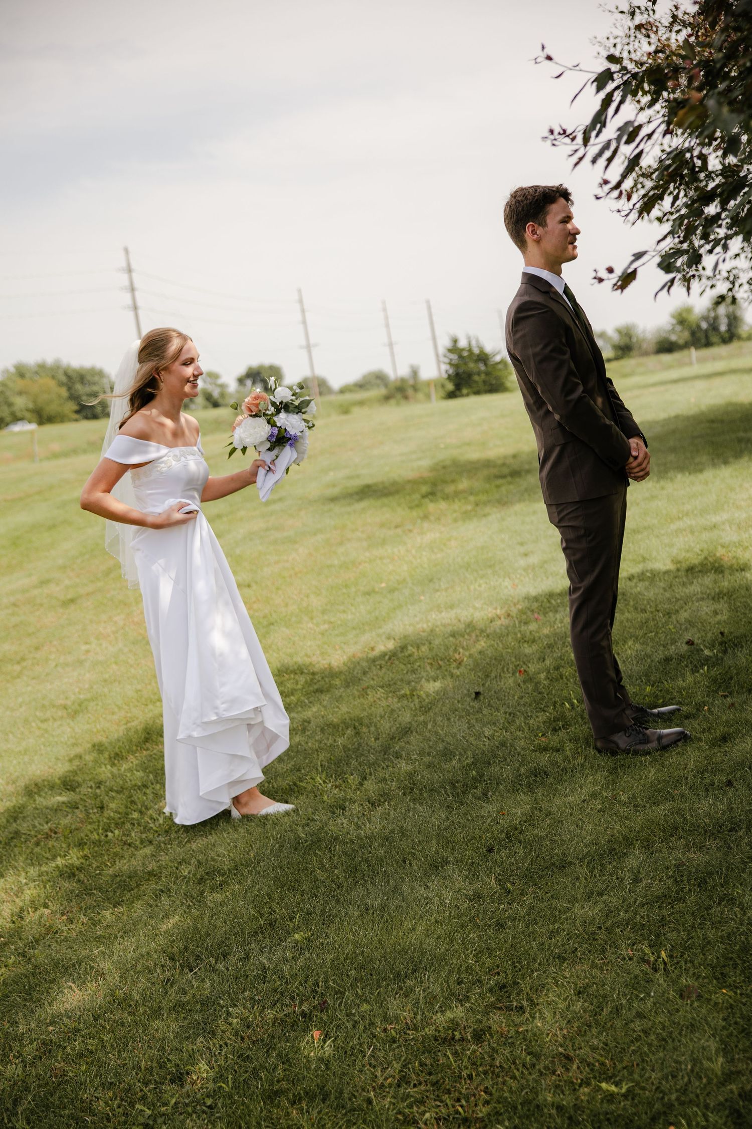 Smiling bride in a white gown with a veil holds her bouquet and reaches toward the groom waiting with his back turned in a grassy field.