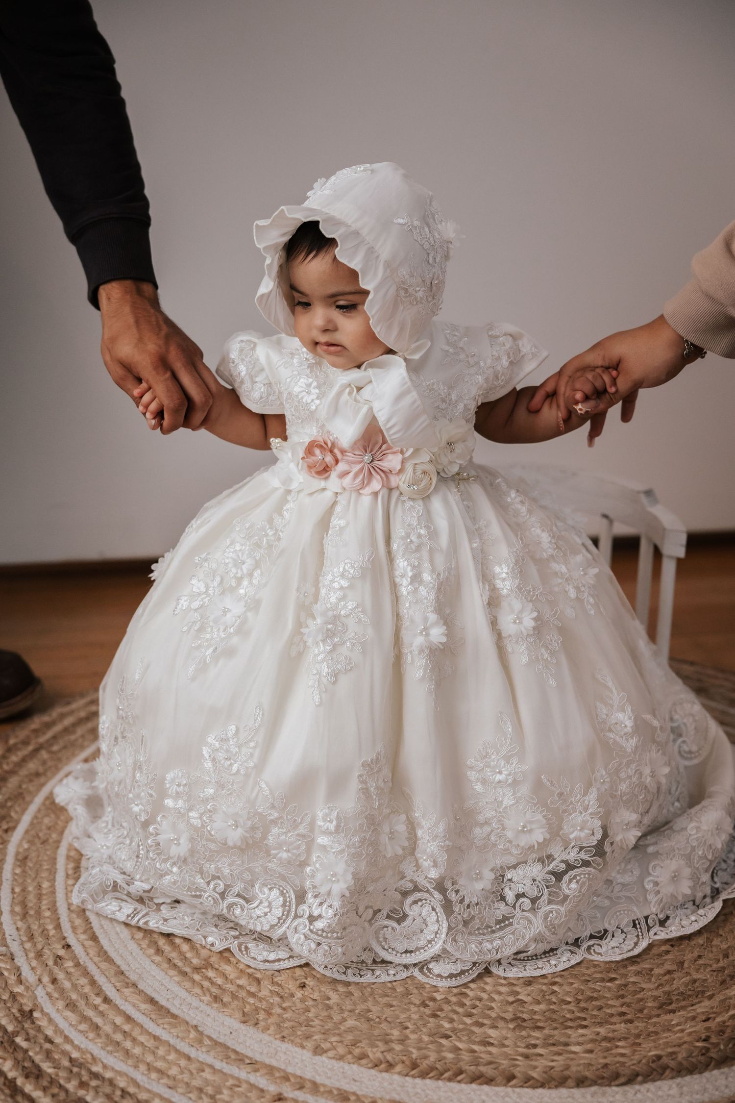 Baby in an ivory lace gown and matching bonnet with pink floral accents stands holding adults' hands on a woven rug.