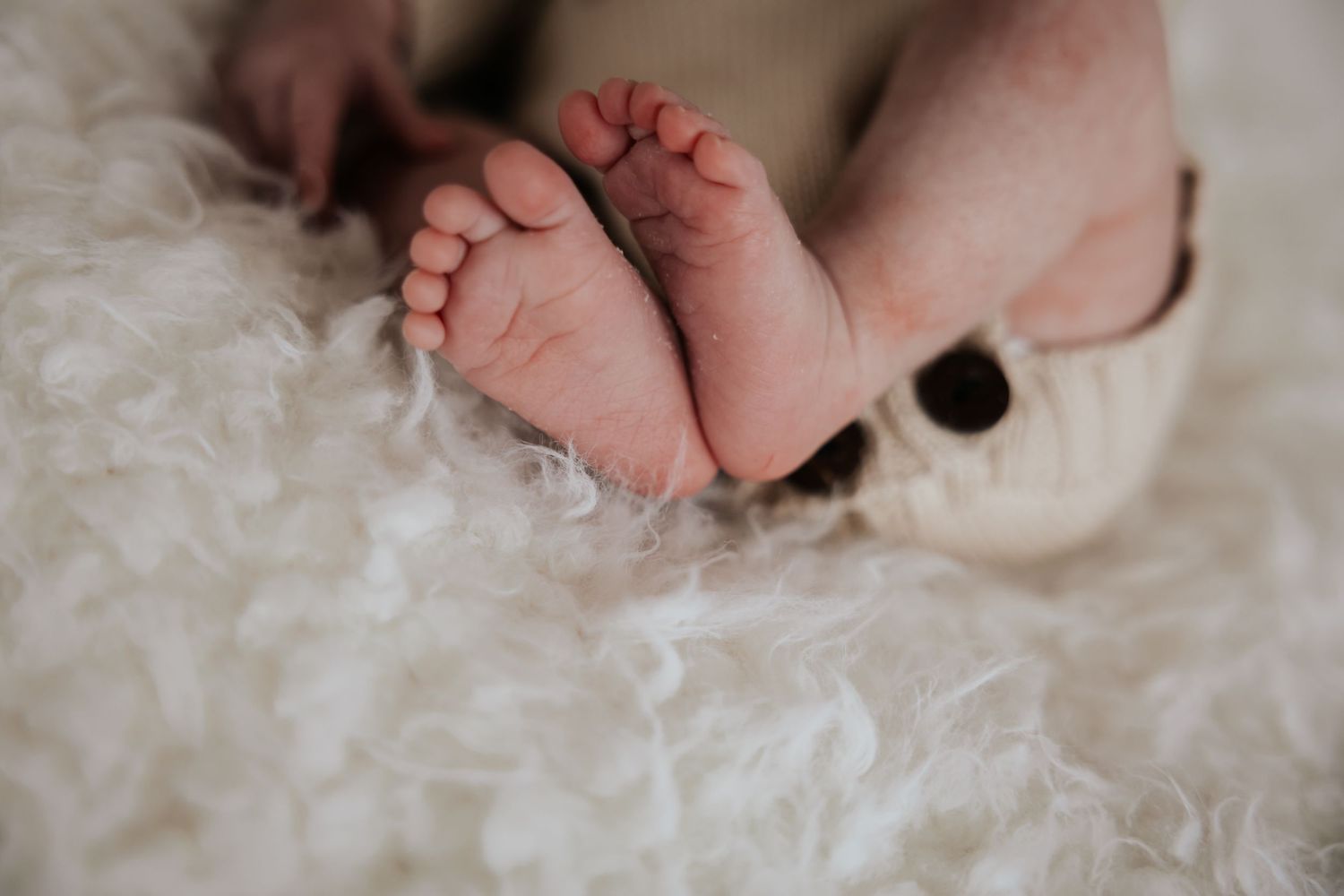 Close-up of tiny newborn baby feet resting on a soft white fluffy blanket beside a cream knit romper.
