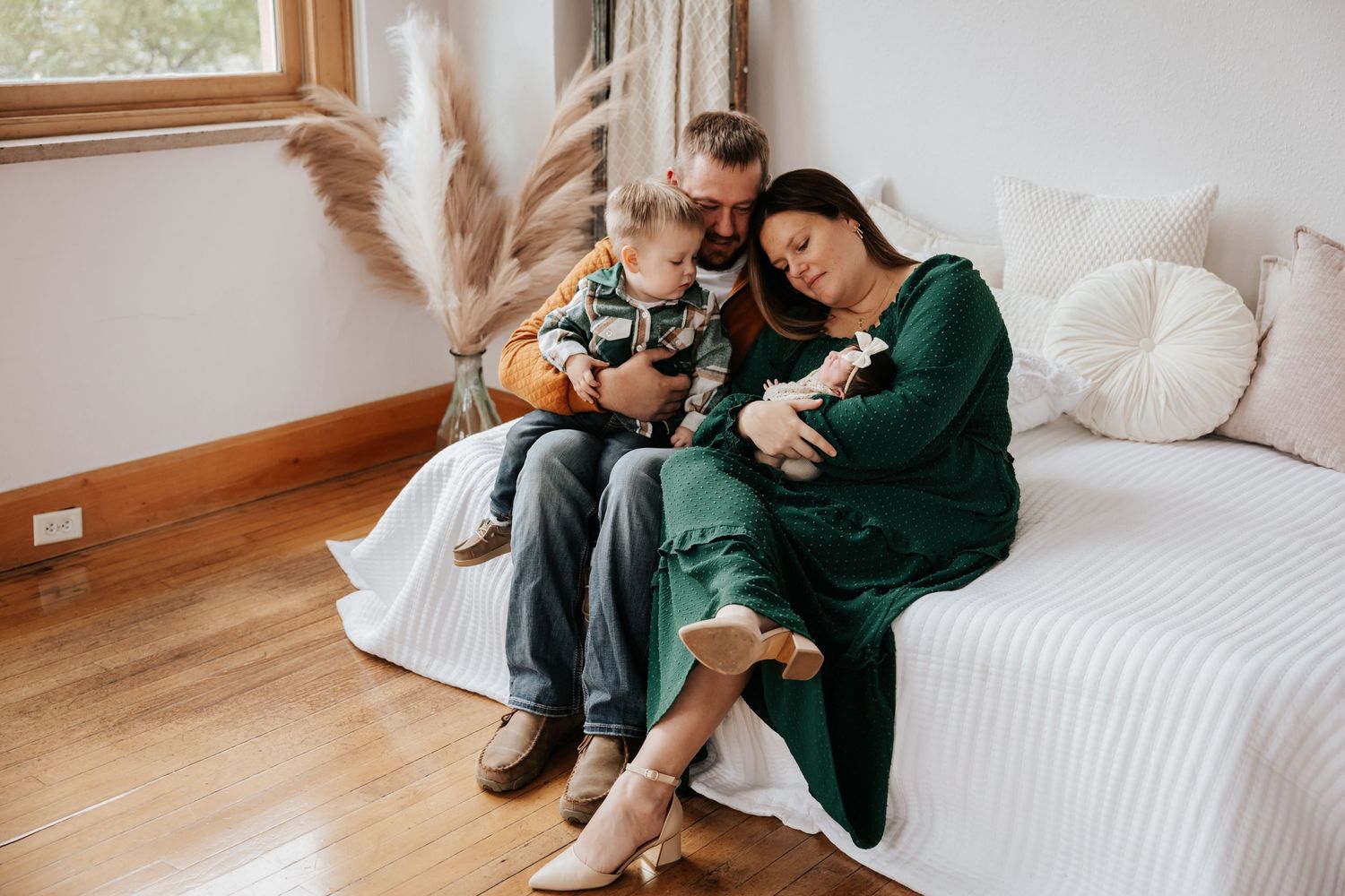 A cozy lifestyle photo of parents and a toddler gazing at a newborn on a white daybed beside dried pampas grass.