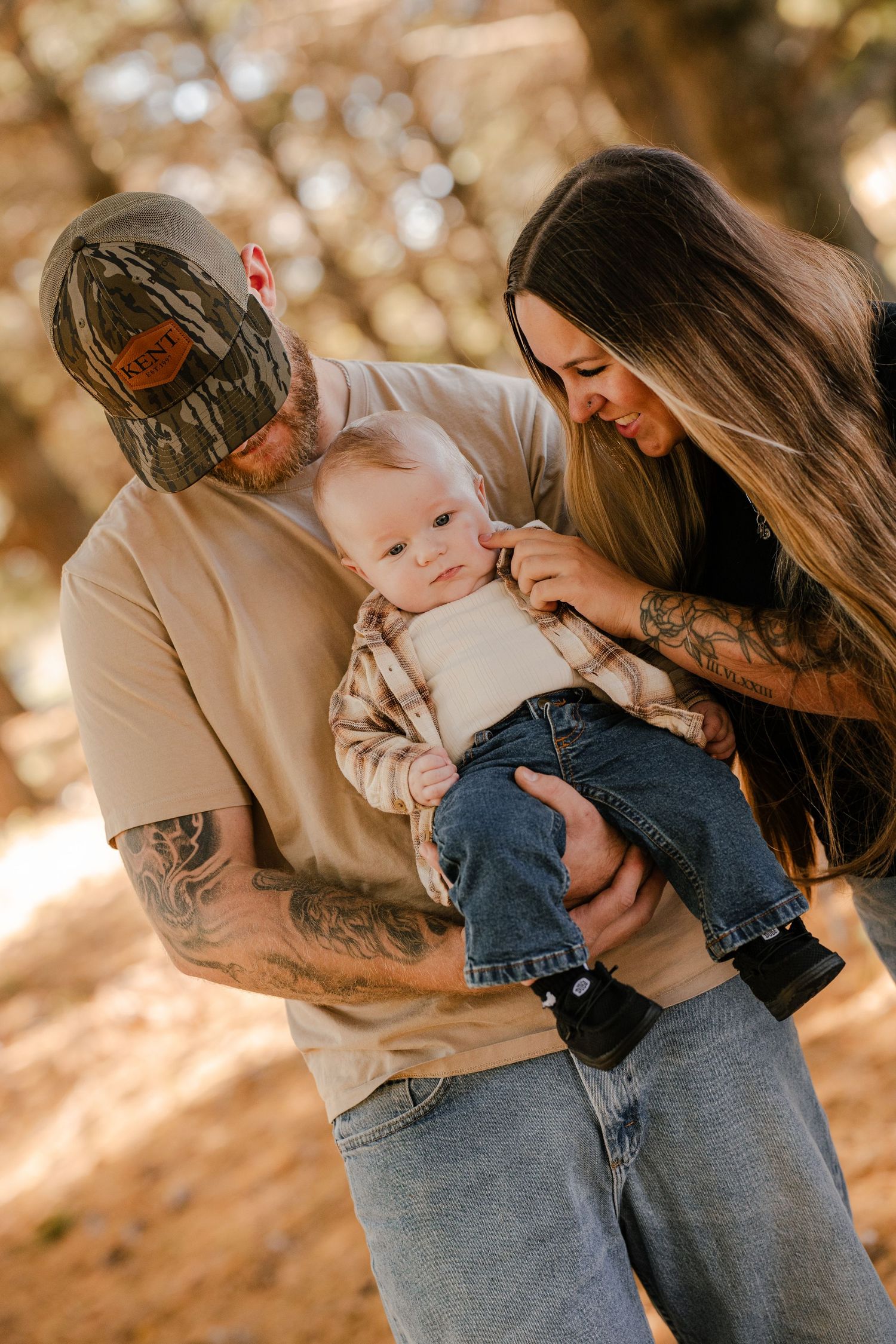 Dad in a camo cap and mom with long hair lovingly look down at their baby in a flannel shirt and jeans in autumn woods.