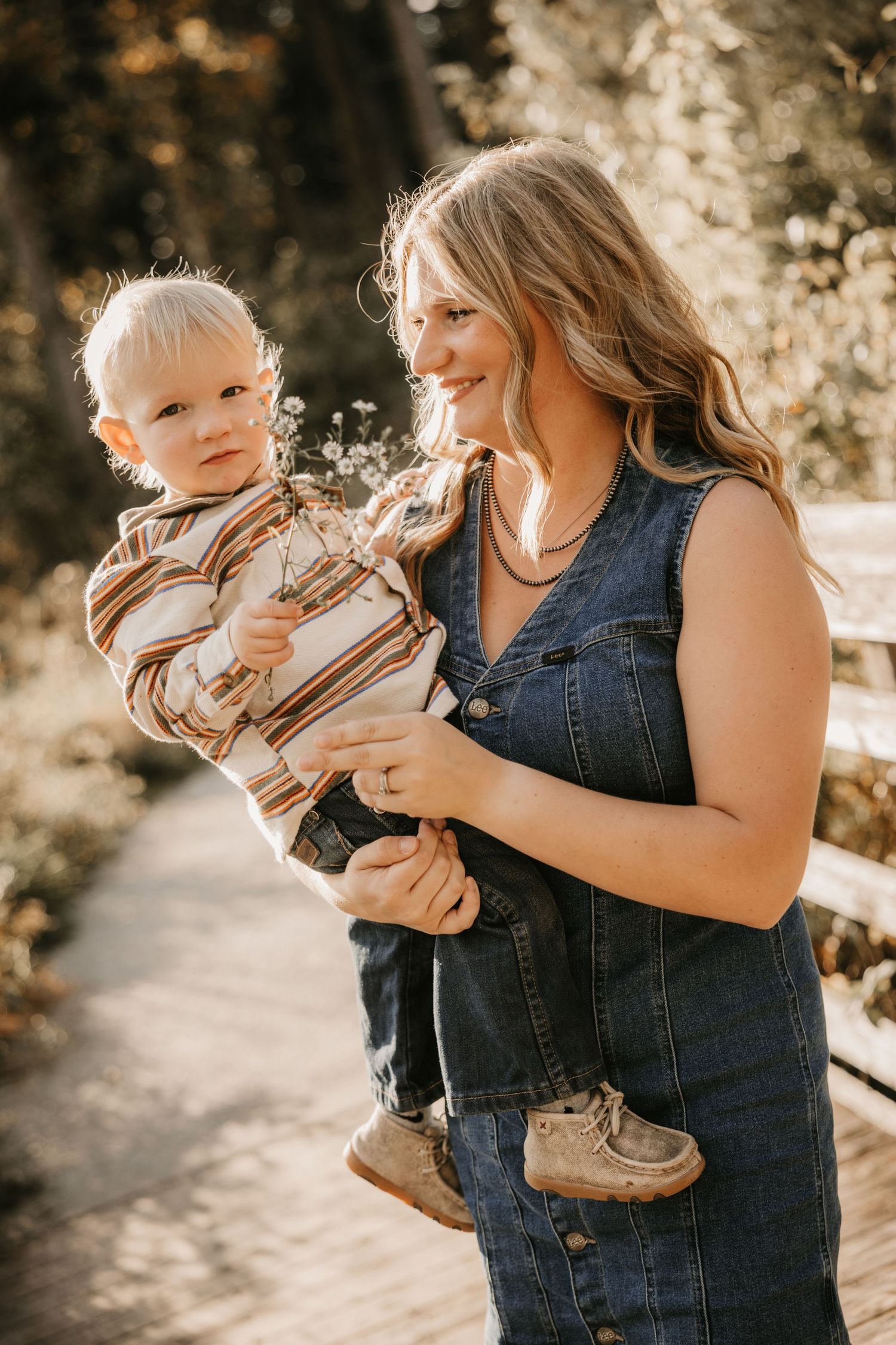 A smiling woman in a denim dress holds a blond toddler in a striped sweater on a sunlit wooden boardwalk.