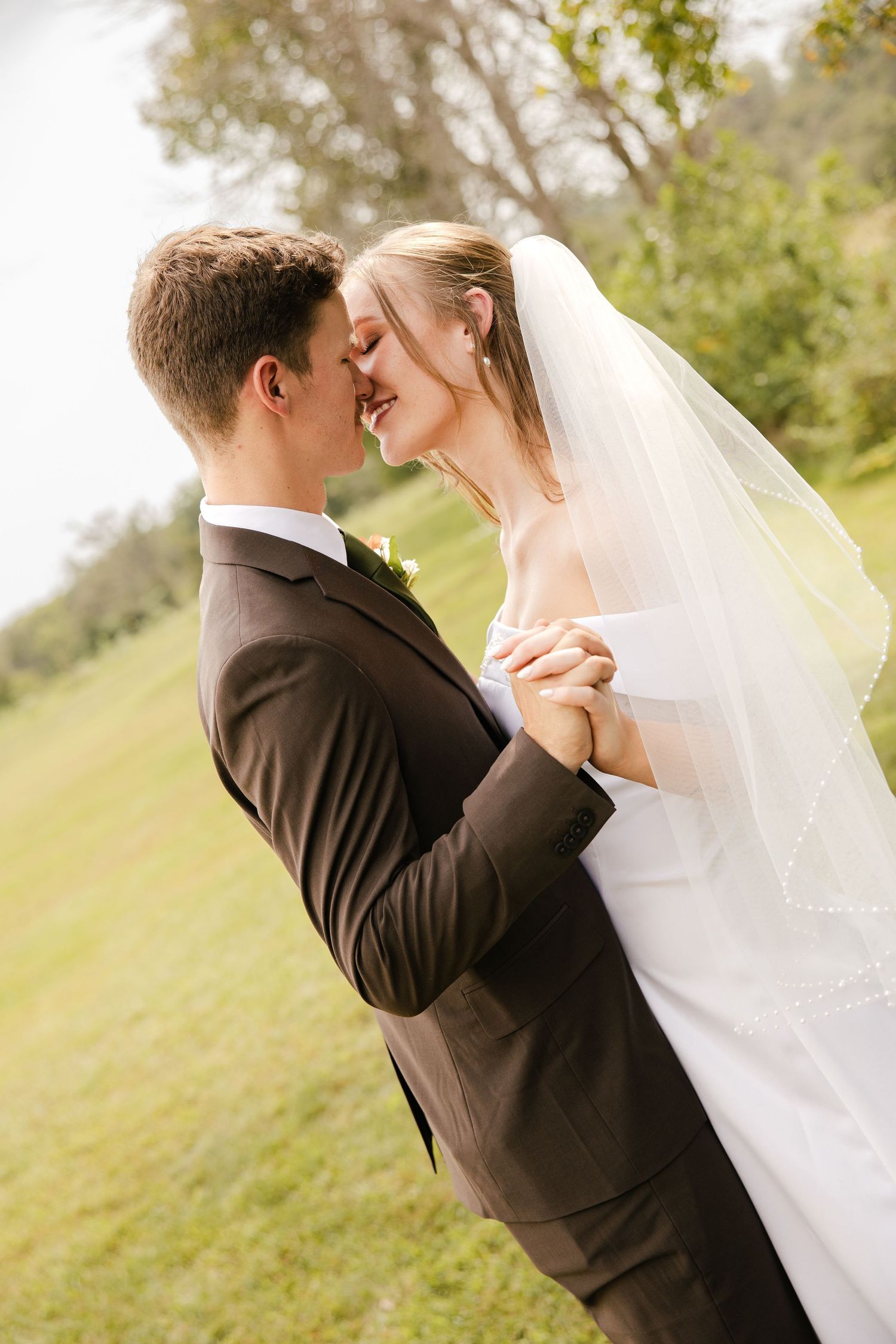 A bride and groom lean in for a tender kiss while holding hands outdoors, she wearing a beaded veil and he a brown suit with boutonniere.