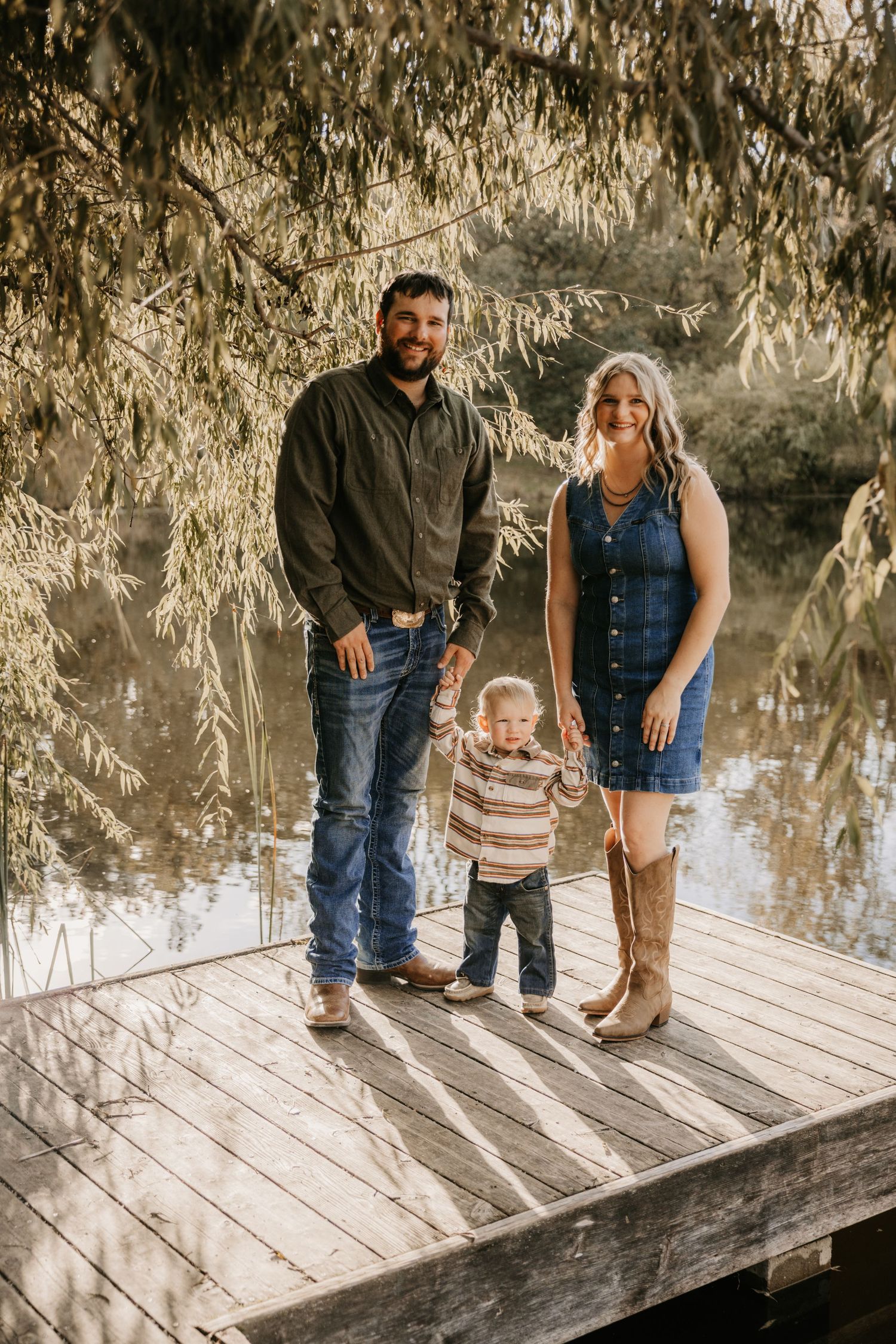 Parents hold their toddler's hands as he stands between them on a wooden dock beside a lake and willow trees.