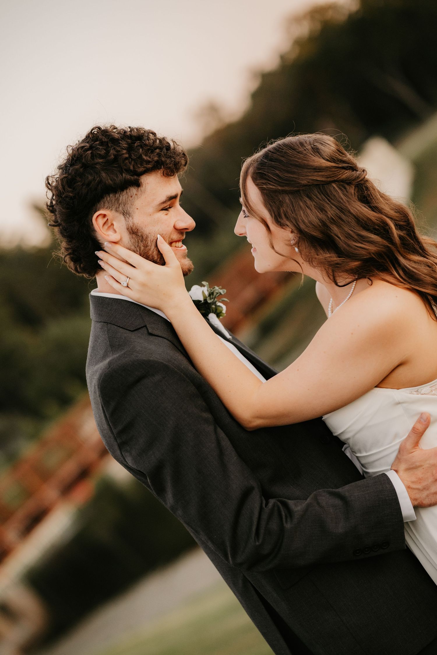 Bride in a white strapless gown touches her groom's cheek as the smiling couple embrace in warm golden evening light.