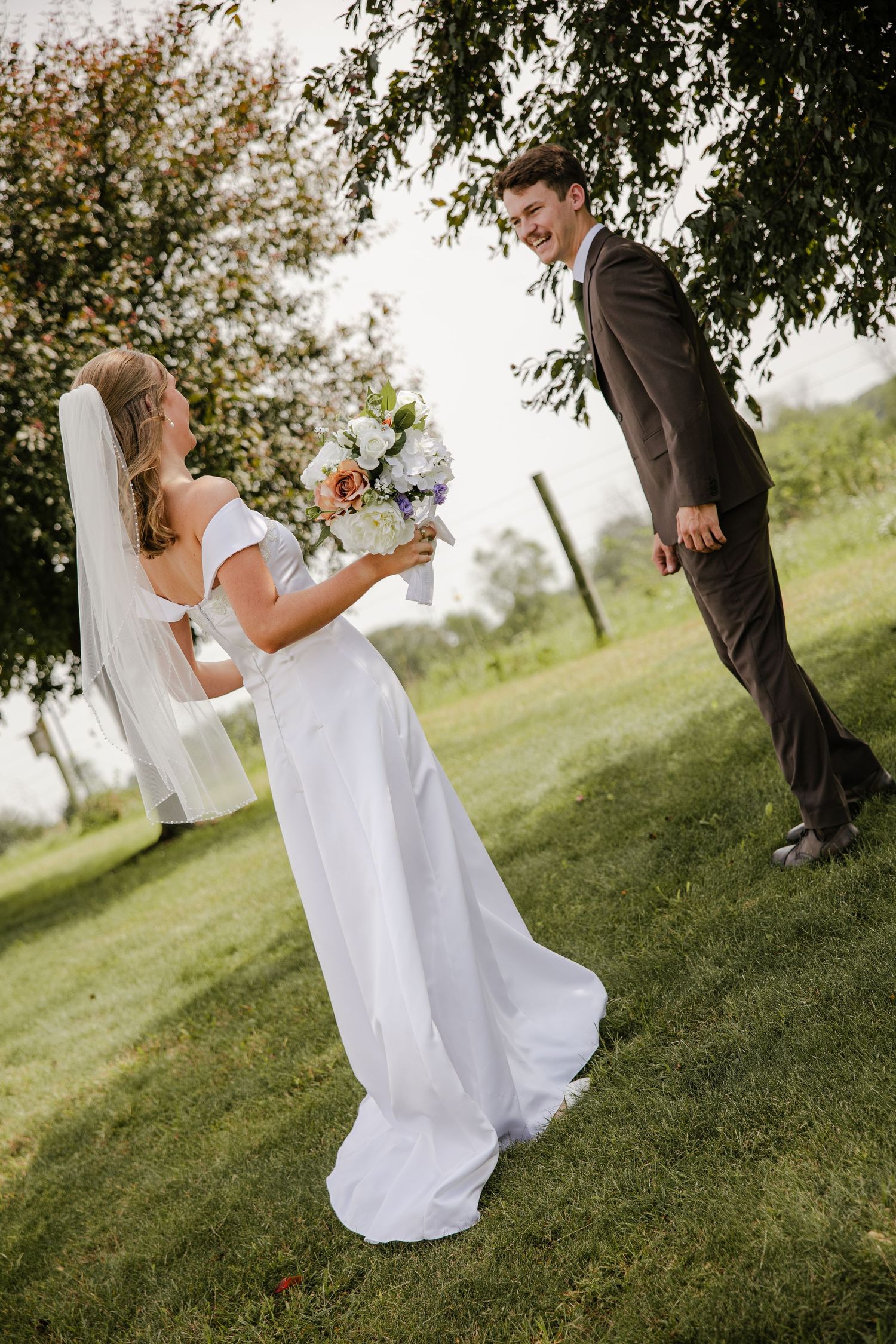 Groom in a brown suit turns and smiles at his veiled bride holding a floral bouquet during their first look under a leafy tree.