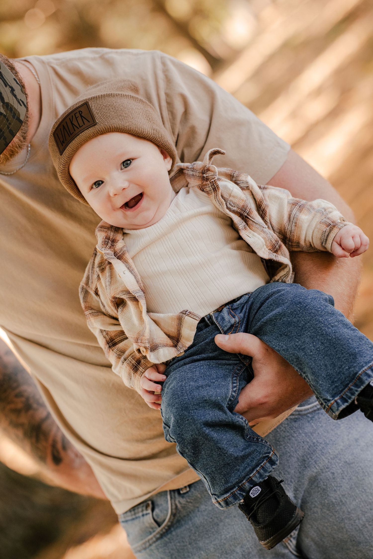 Happy baby in a WALKER beanie and flannel shirt is cradled in a parent's arms among sunlit trees.