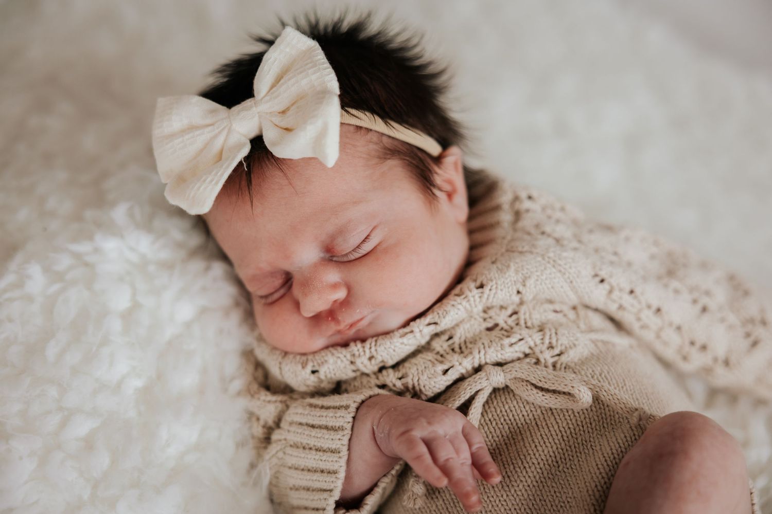 Sleeping newborn baby girl wearing a cream bow headband and knit romper resting on a soft fluffy white blanket.