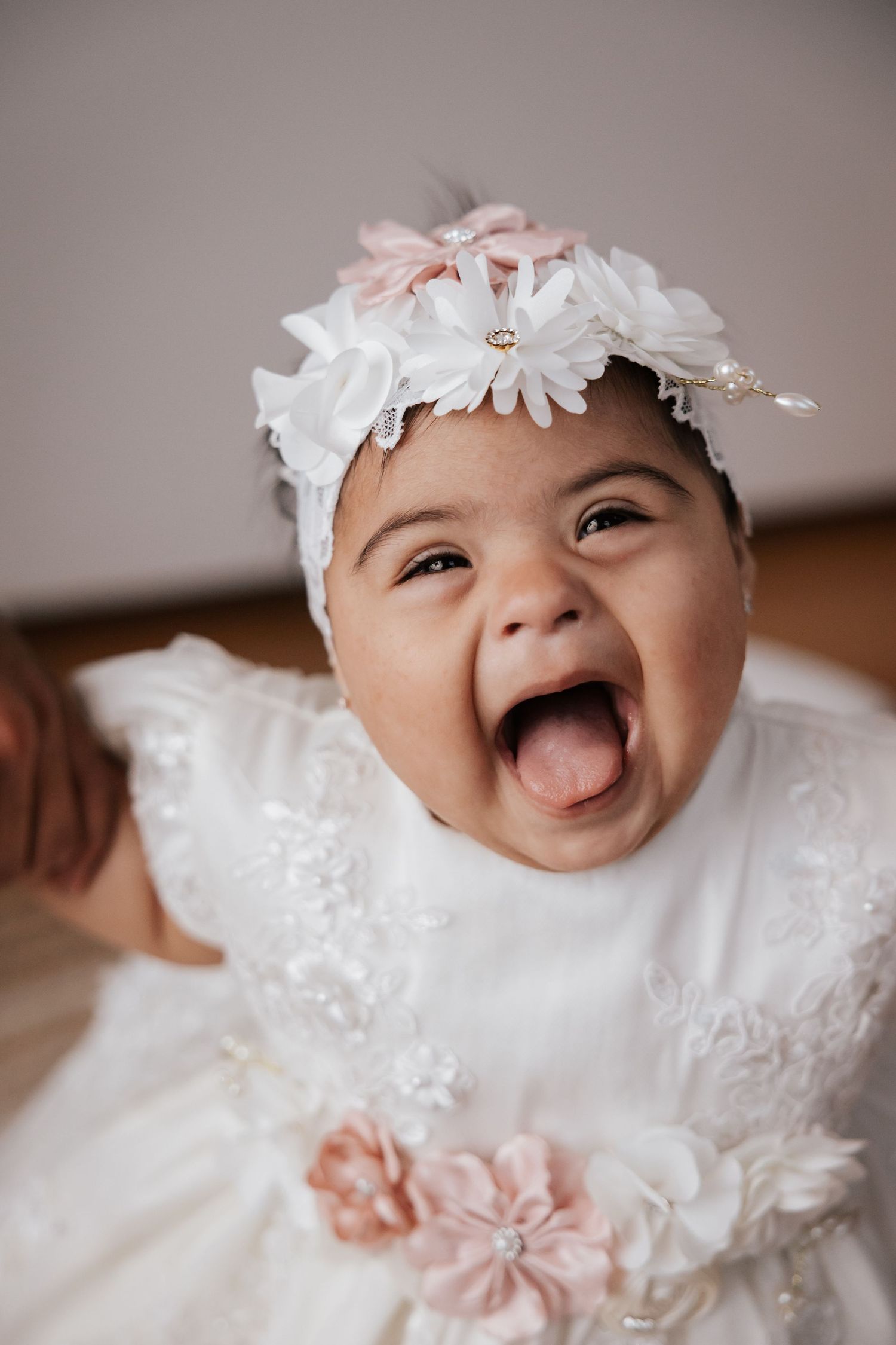 Laughing baby girl in a white lace dress and floral headband with pink and white flowers, mouth wide open in joy.