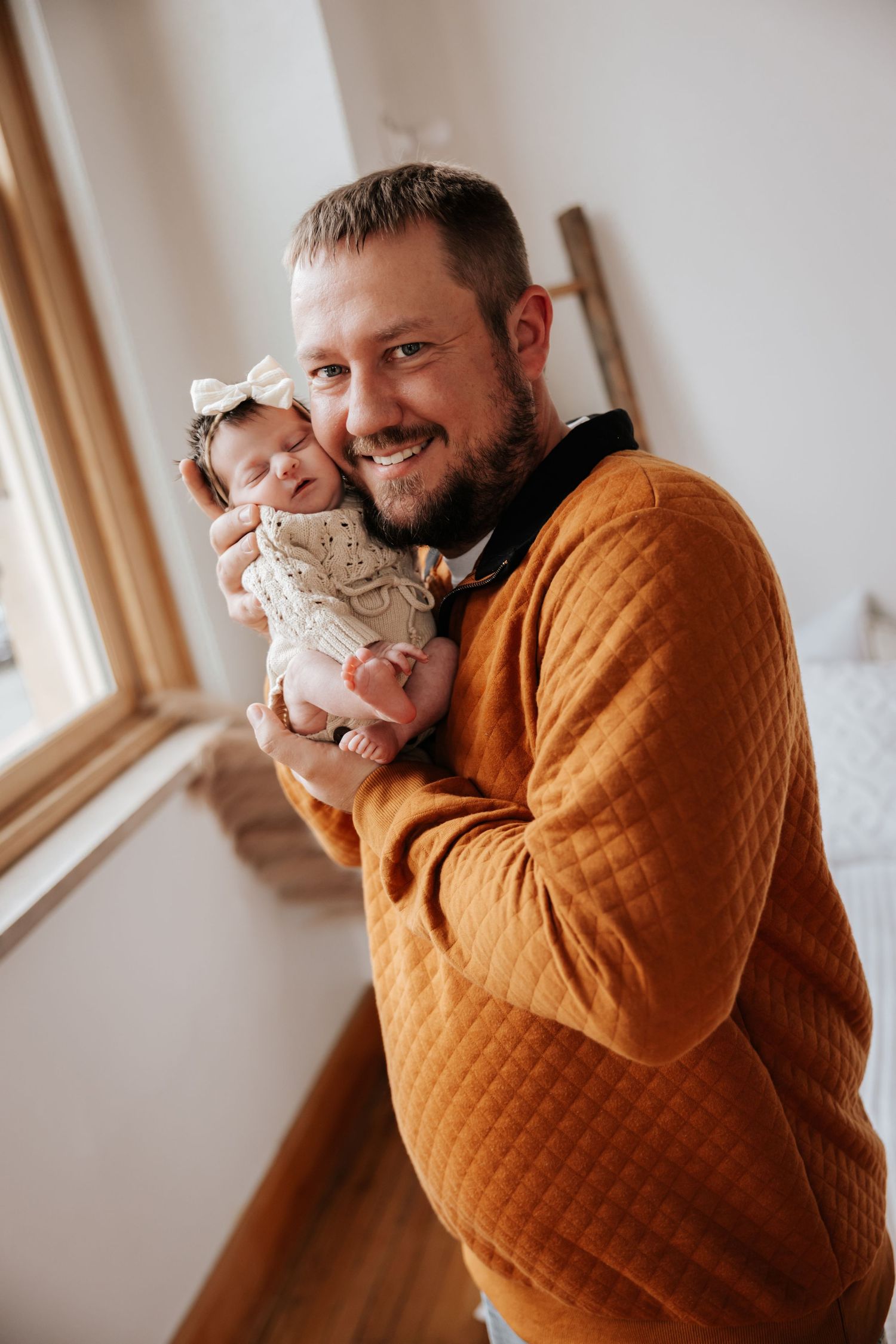 Smiling dad in an orange quilted sweater holds his newborn baby girl in a cream knit outfit and bow by a window.