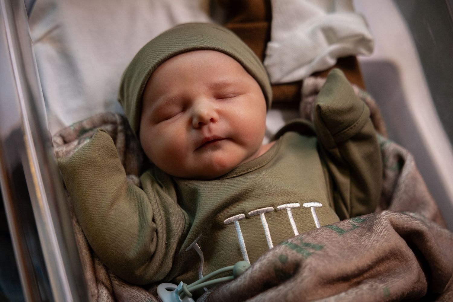 A sleeping newborn baby in an olive green outfit and matching beanie rests in a hospital bassinet wrapped in a soft blanket.
