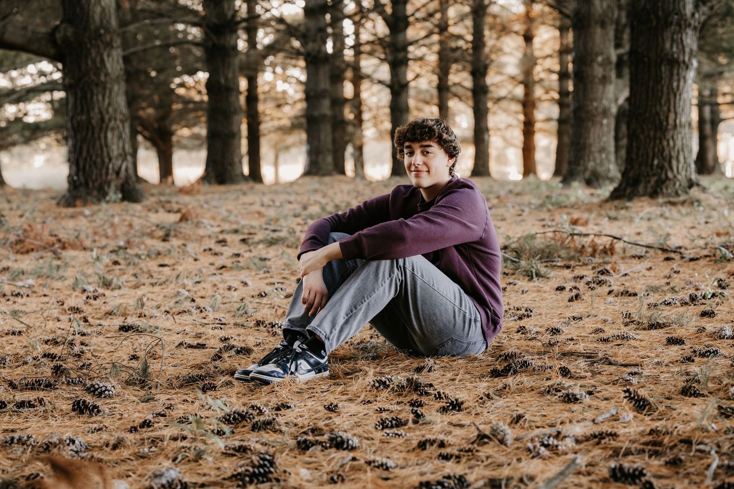 Young man in a maroon sweatshirt and gray jeans sits on a carpet of pine needles among tall trees, smiling softly at the camera.