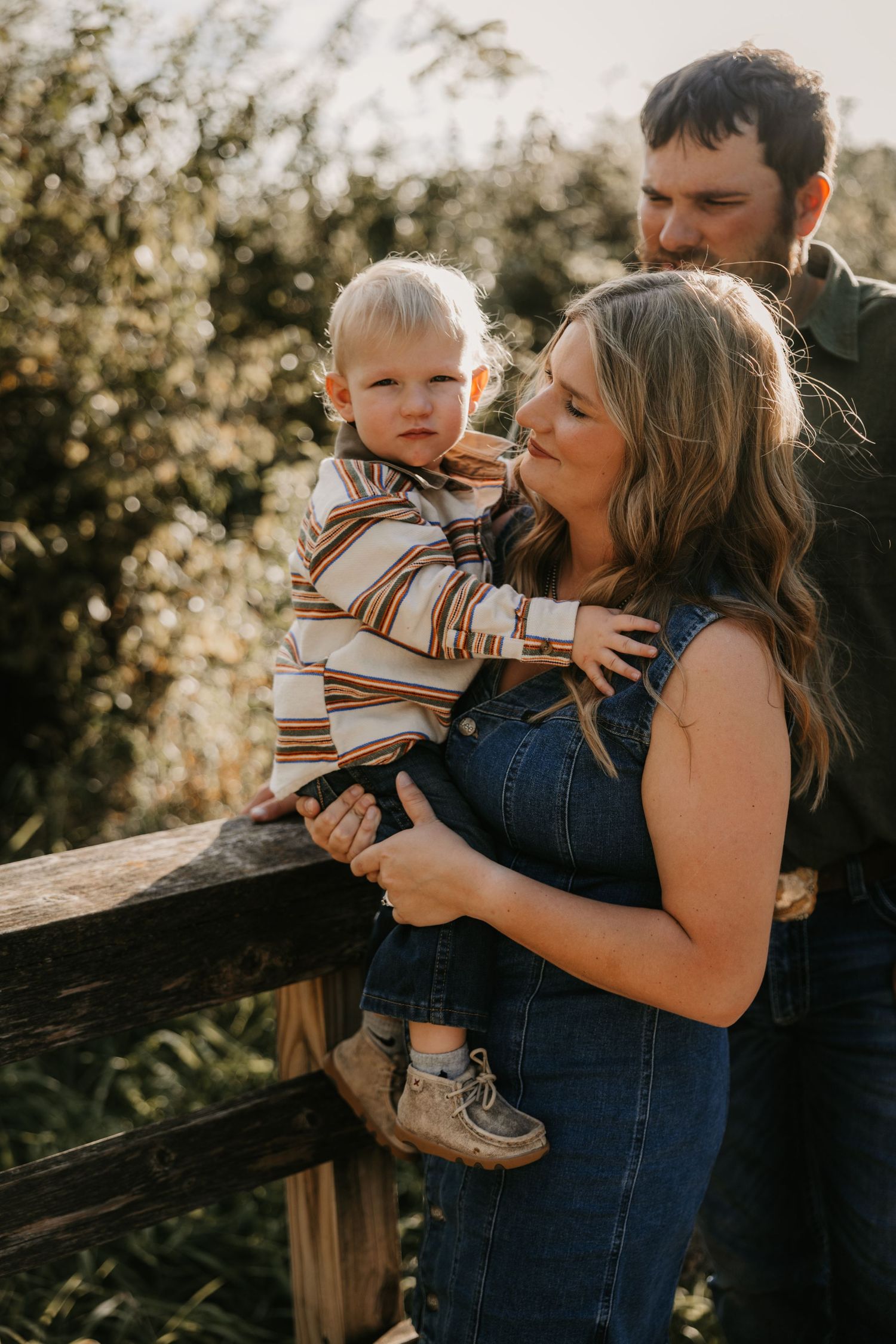A mother in a denim dress holds her toddler son against a wooden fence while the father stands close behind during a golden hour portrait.