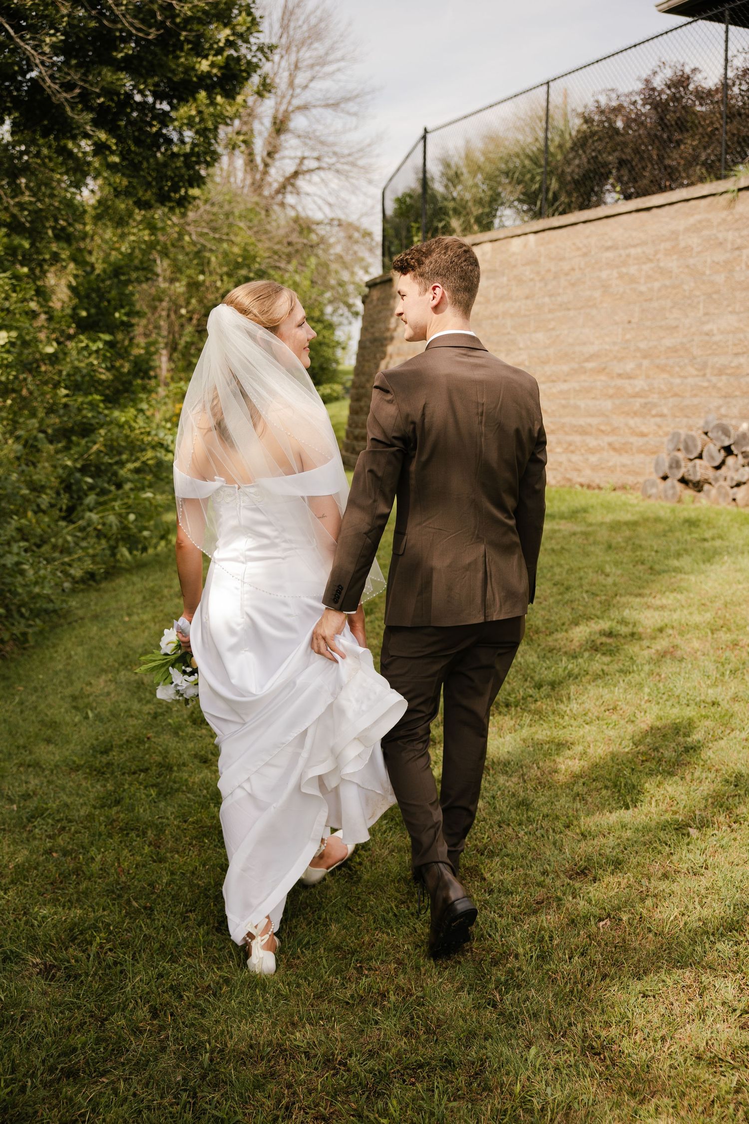 Bride and groom walk hand in hand across a green lawn beside a stone wall on their wedding day.