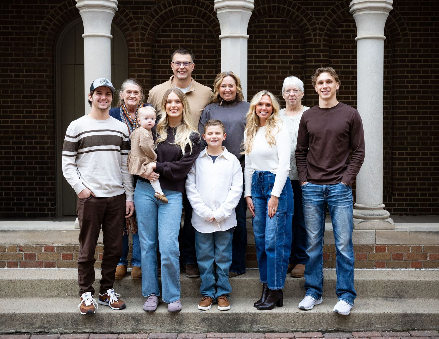 Large family of eleven, from a toddler to grandparents, posing together on stone steps in front of a brick building with white columns.